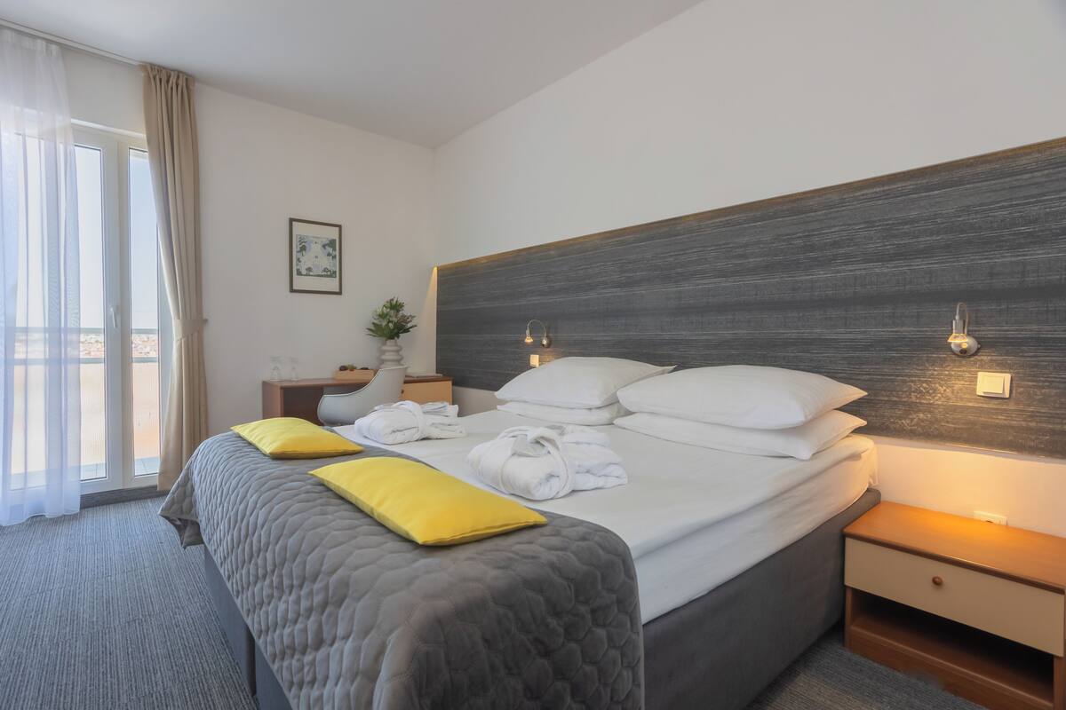 Habitación de hotel con una cama grande, una mesita de noche, una planta en maceta y una ventana con cortinas.