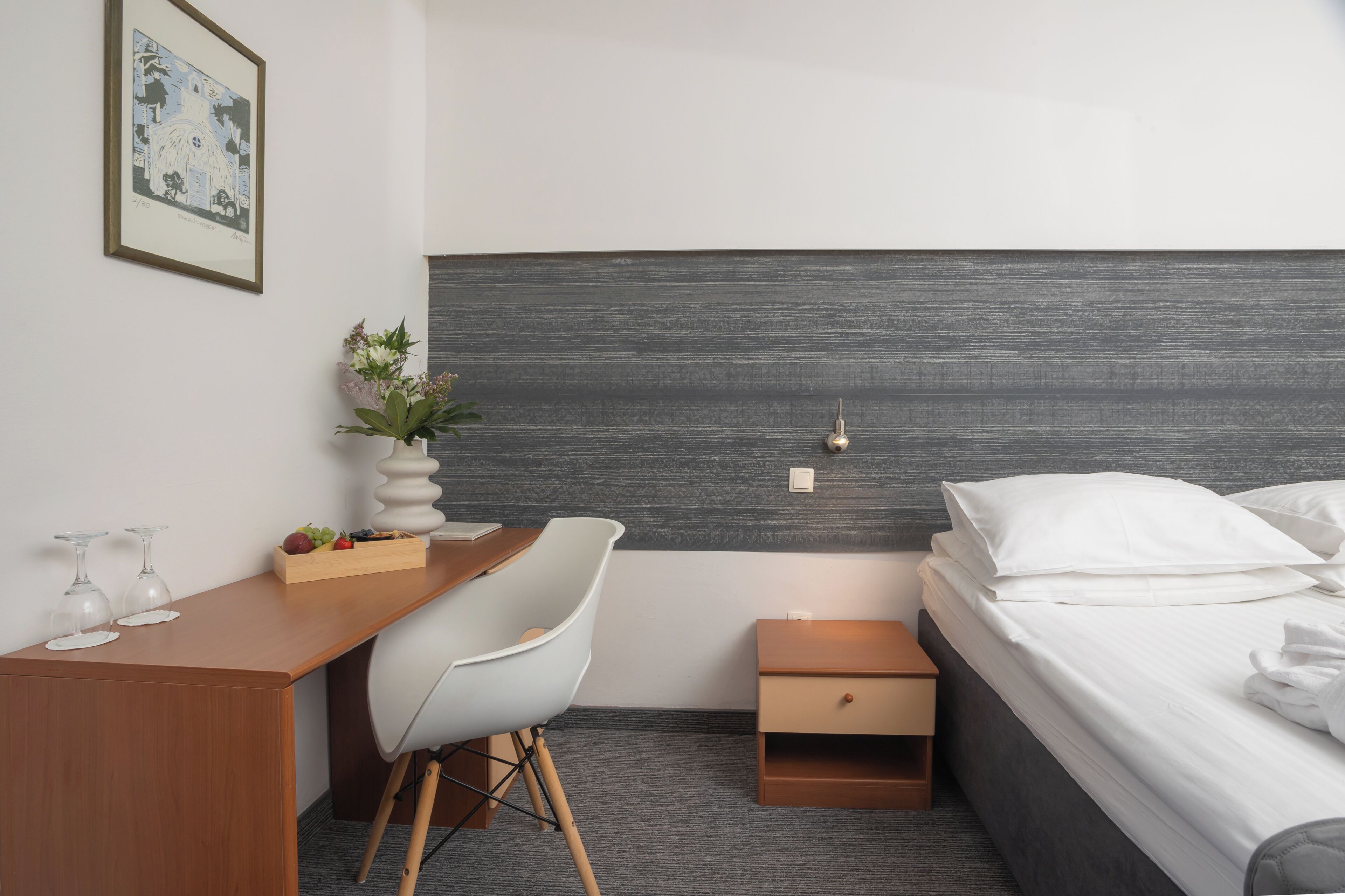 A hotel room with a bed, a desk, a chair, a small bedside table, a nightstand, a wall-mounted light, and a framed picture on the wall.