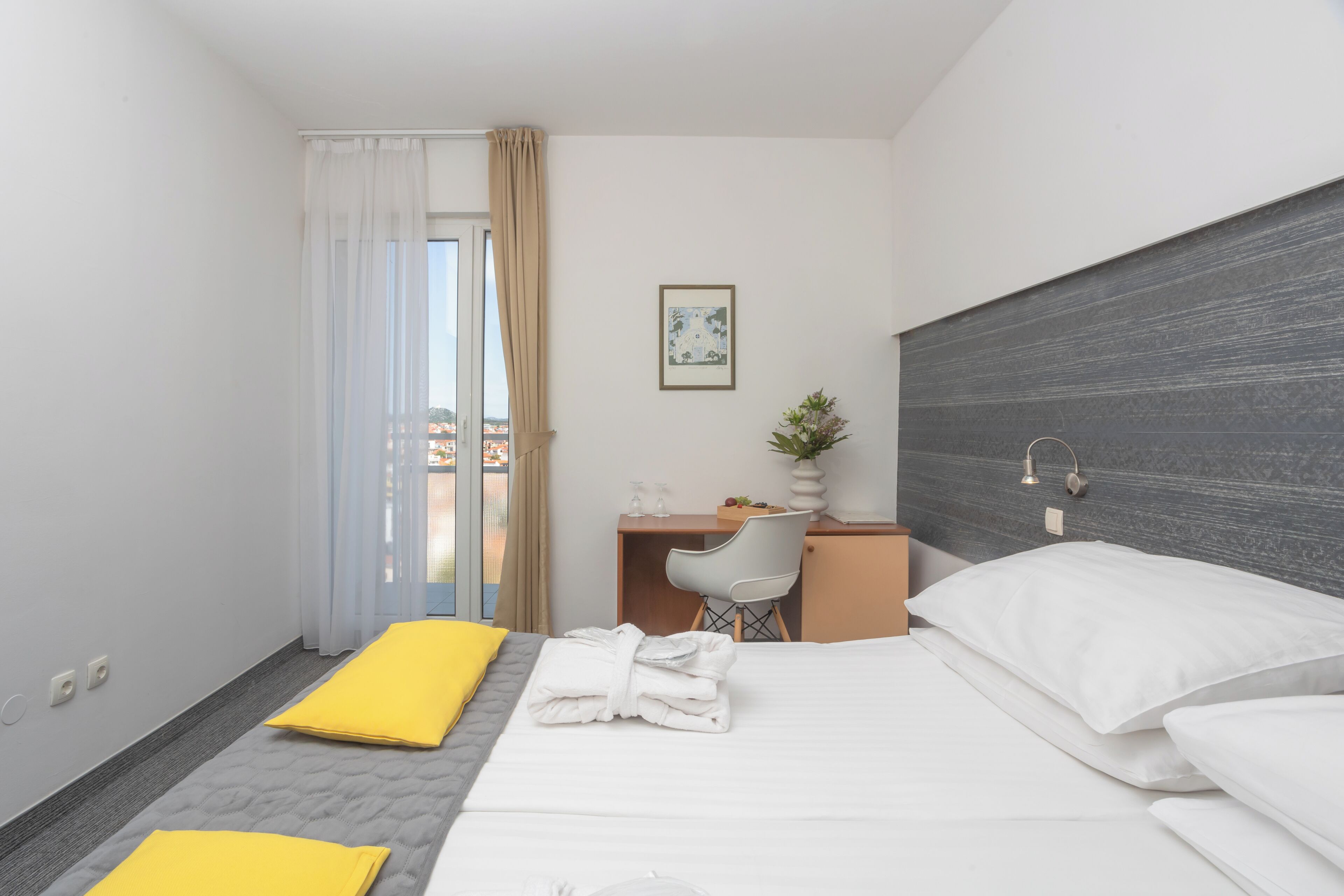 A hotel room with a bed, a desk, a chair, a small table, a potted plant, and a view through a window.