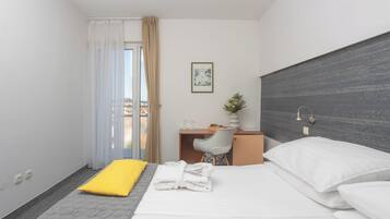 A hotel room with a bed, a desk, a chair, a small table, a potted plant, and a view through a window.