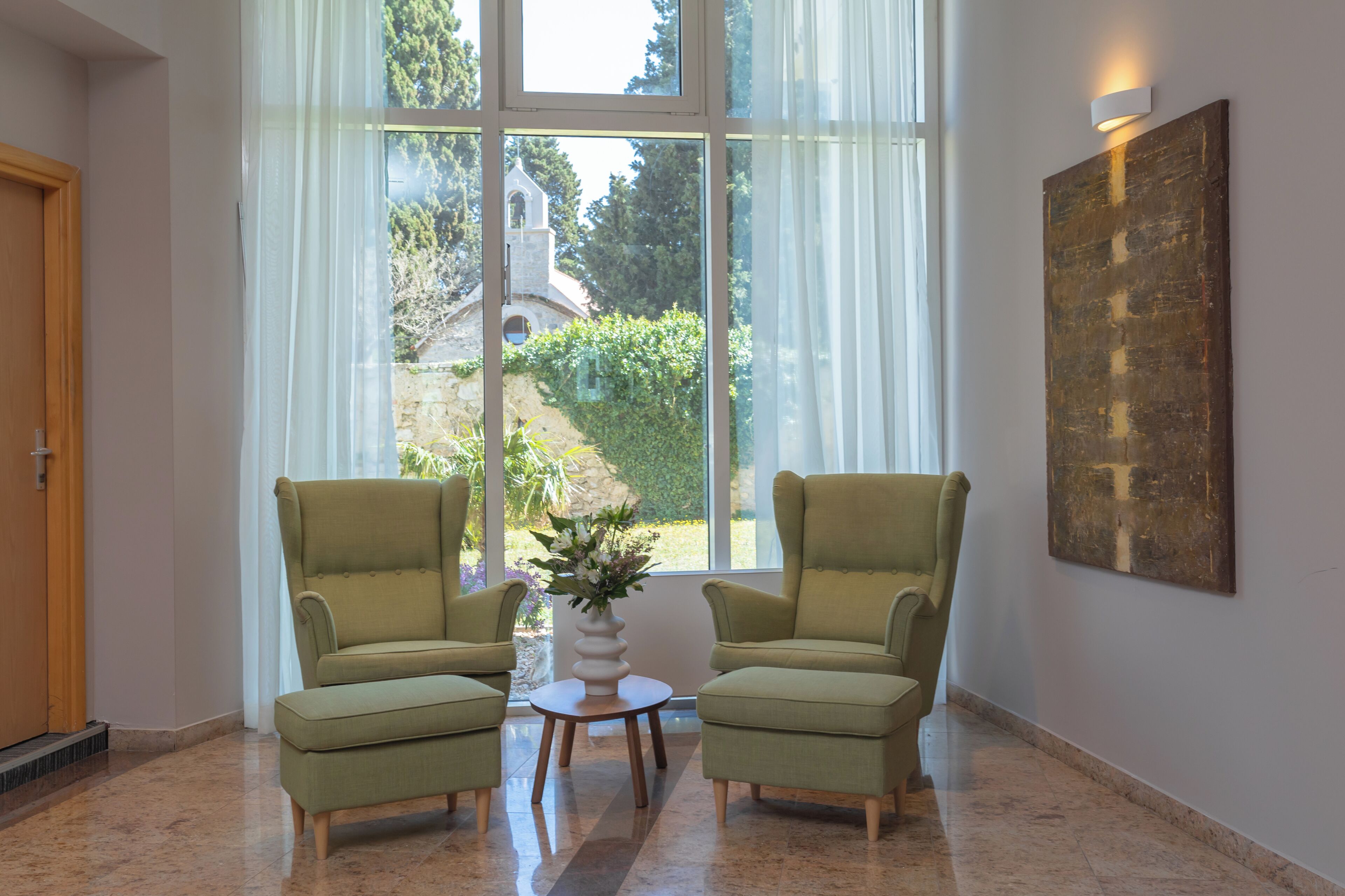 A room with two armchairs, a small table with a vase, a large window with a view of greenery and a building, and a wall-mounted light fixture.