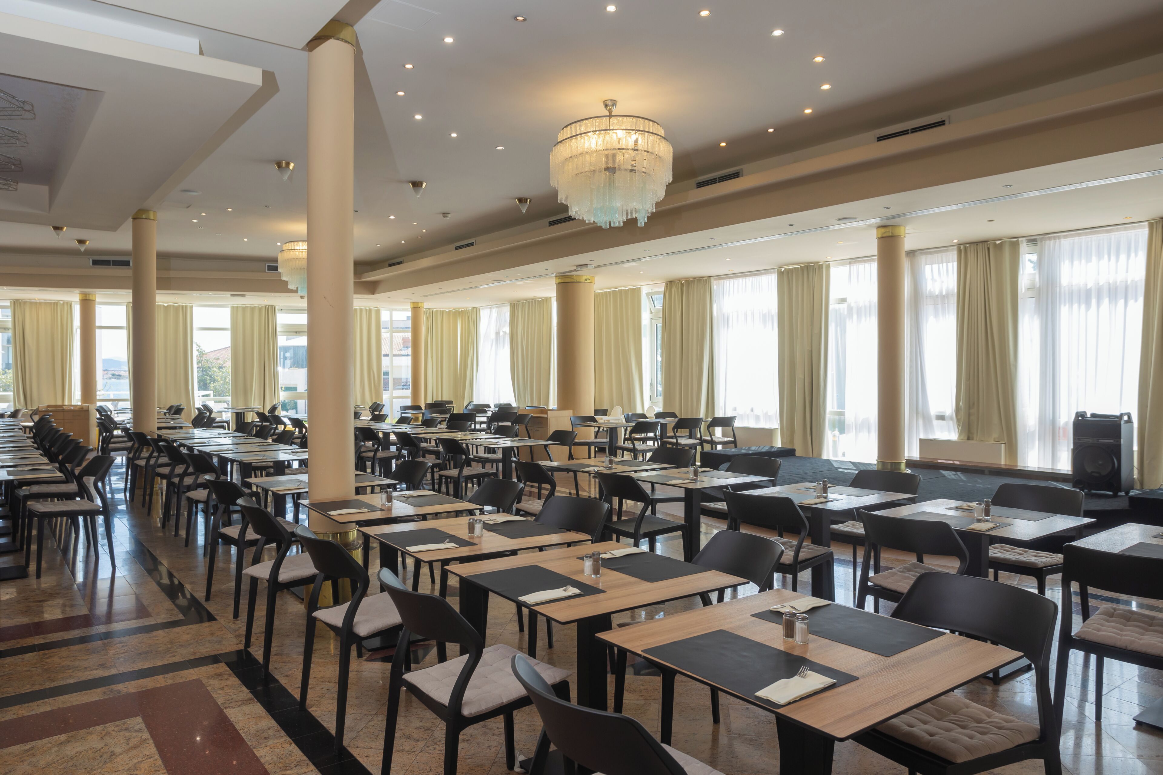 A spacious dining area with multiple tables and chairs, large windows with curtains, and a chandelier.