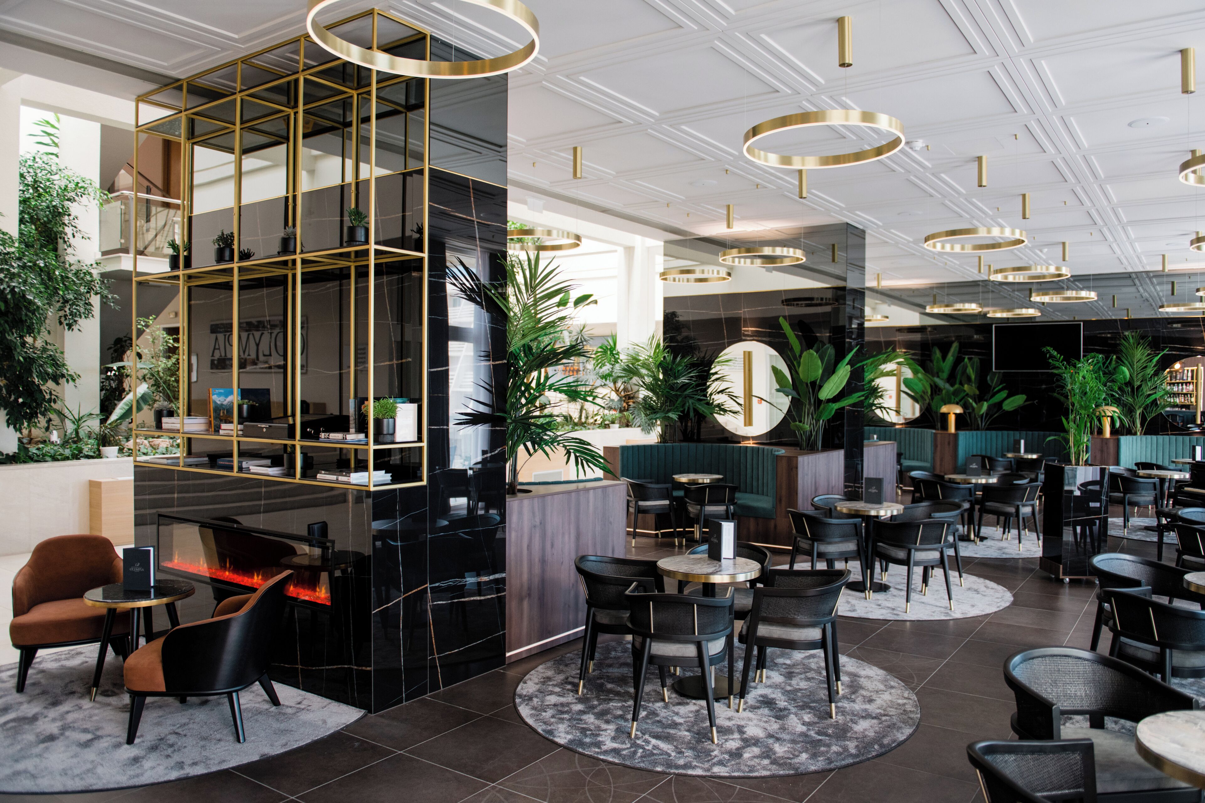 Un moderno vestíbulo de hotel con un bar central, varias zonas de estar y una gran variedad de plantas.