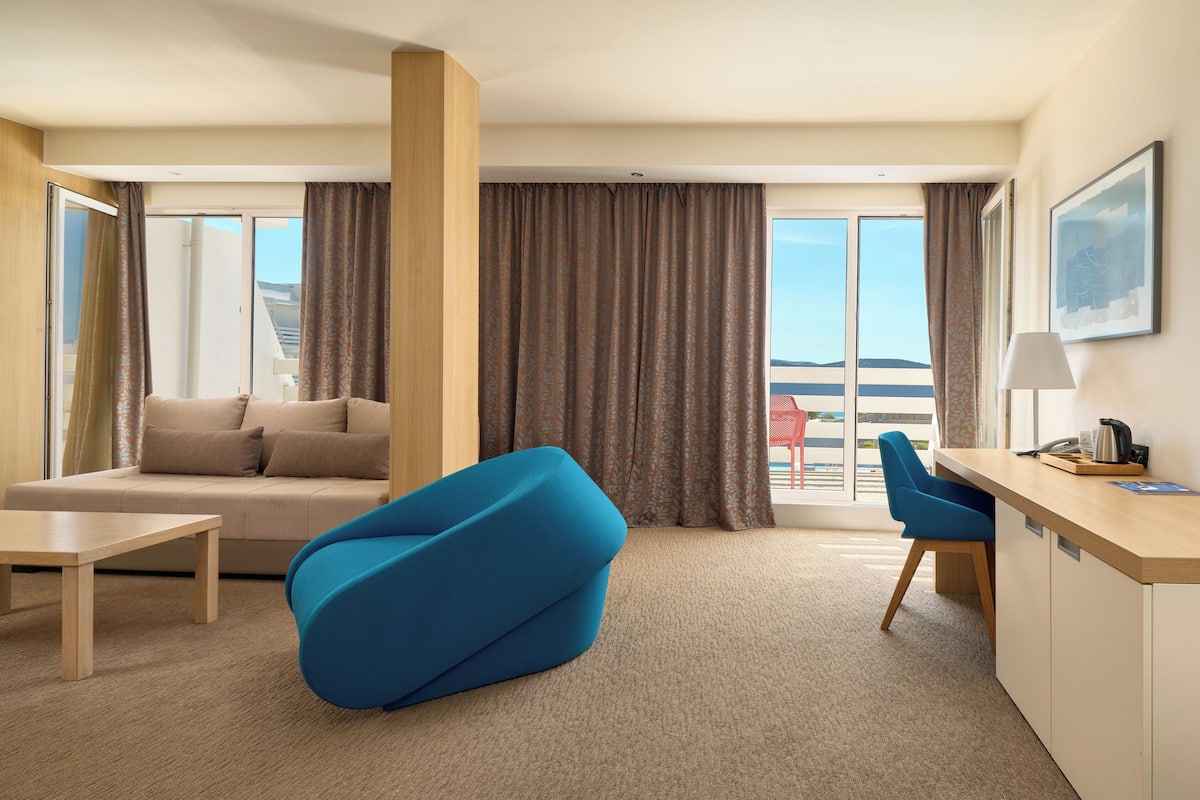 Ein modernes Hotelzimmer mit einer Couch, einem blauen Sessel, einem Schreibtisch und Meerblick durch große Fenster.