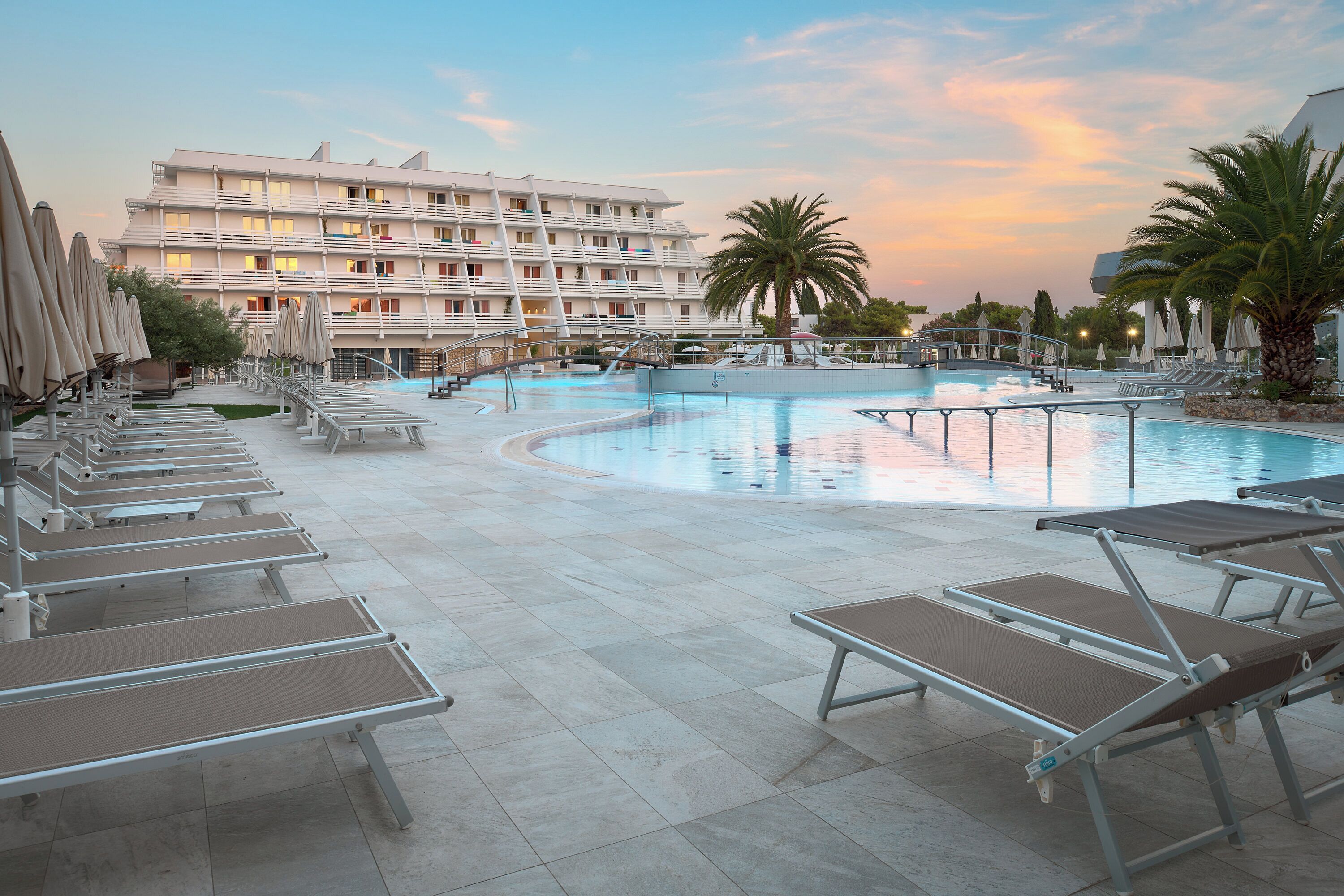 A hotel pool area with sun loungers, a large pool, and a multi-story building in the background.