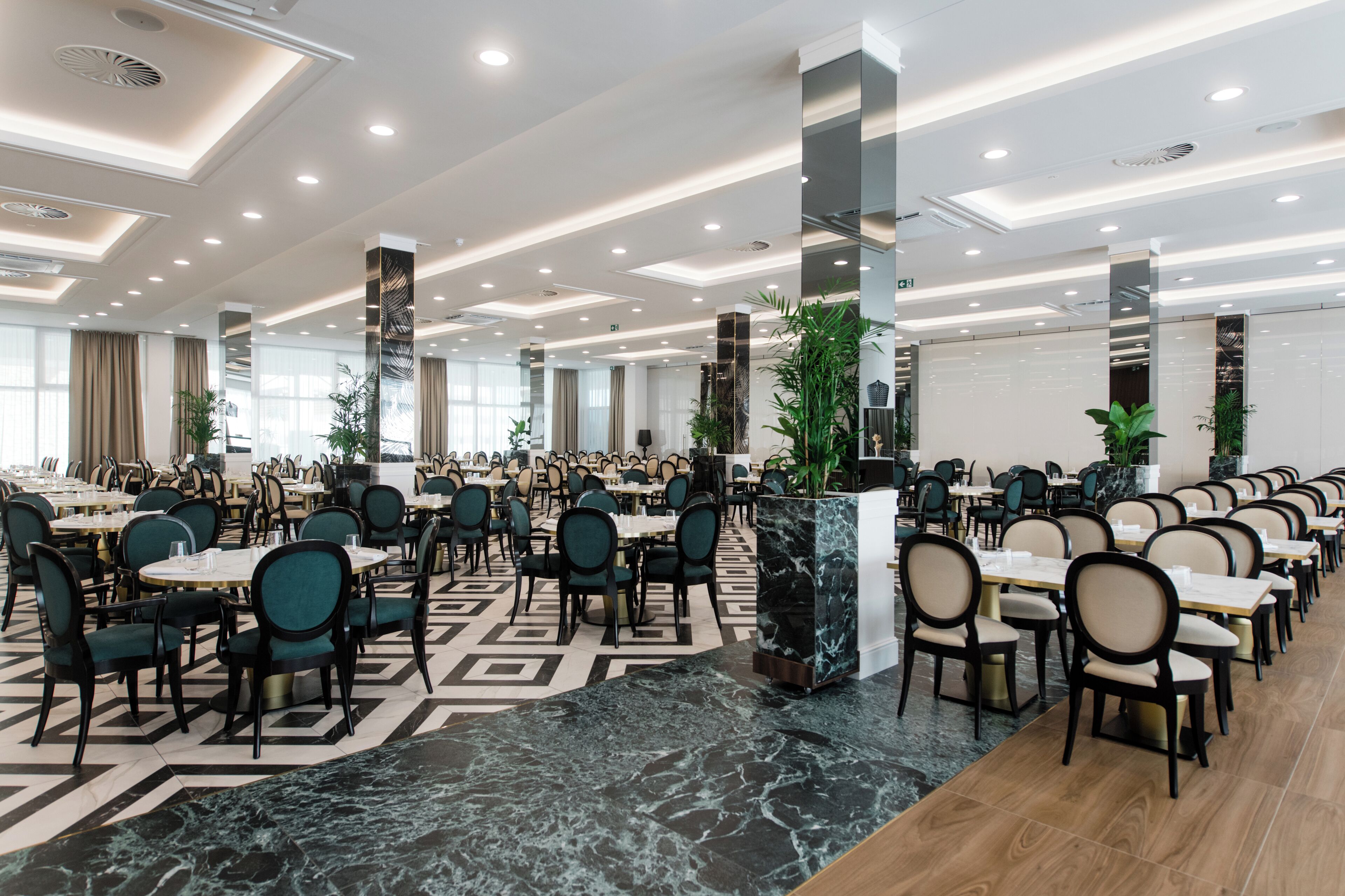 A spacious dining area with multiple round tables and chairs, large pillars, and a patterned floor.