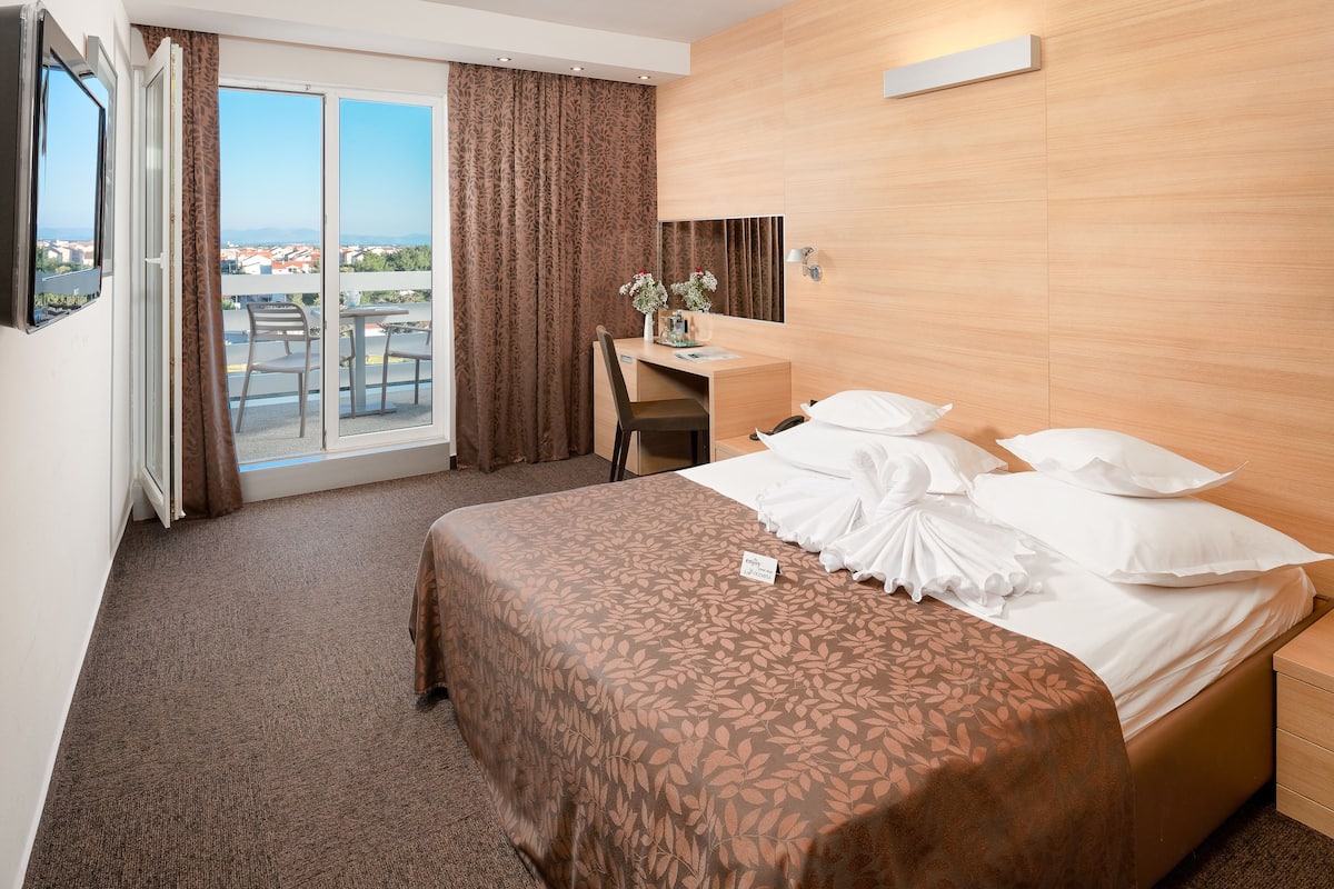 Ein Hotelzimmer mit Bett, Schreibtisch, Stuhl, Fernseher und einem Balkon mit Blick auf Gebäude.