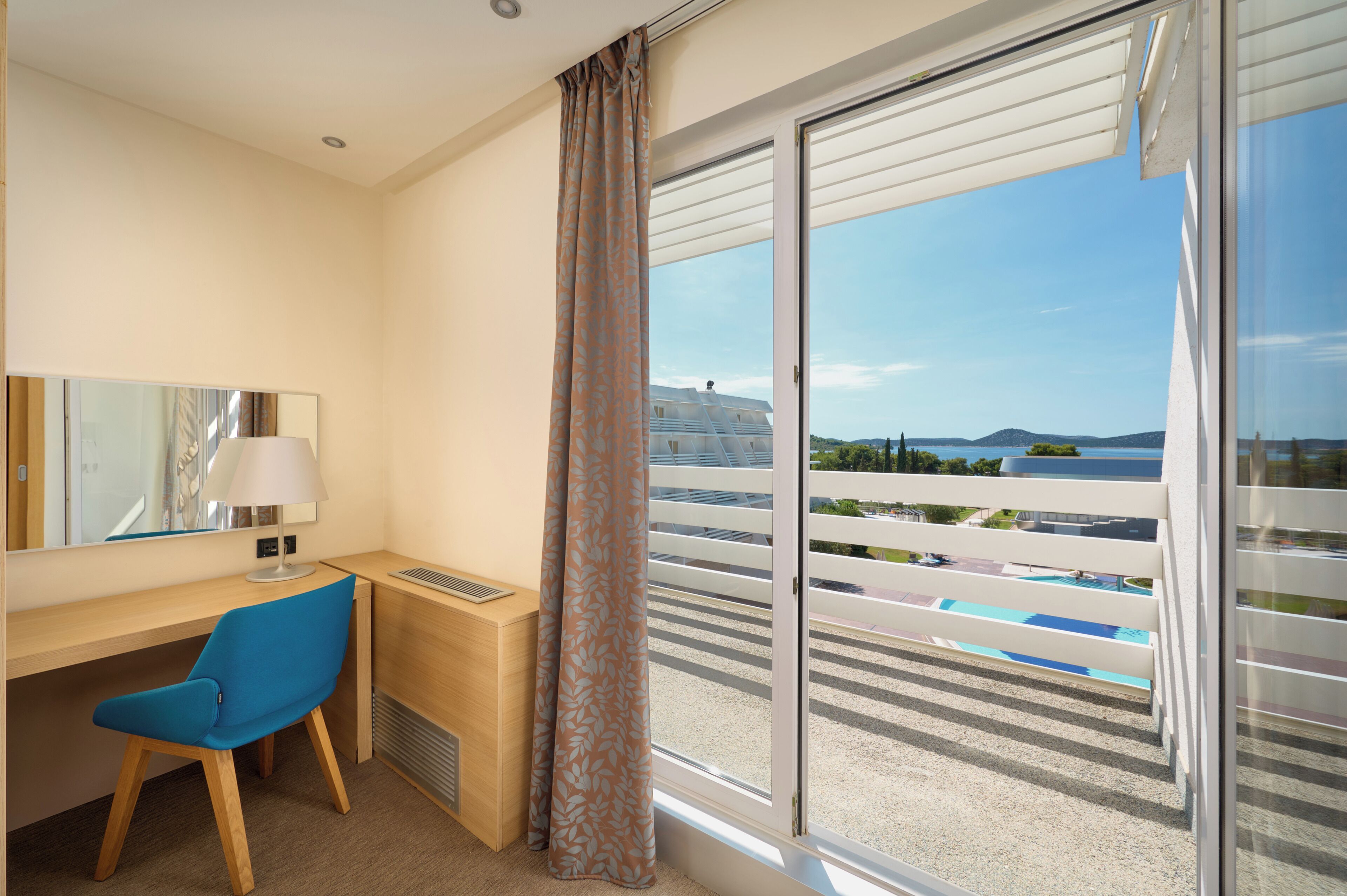 Ett hotellrum med ett skrivbord, en stol och ett stort fönster med utsikt över en pool och byggnader.