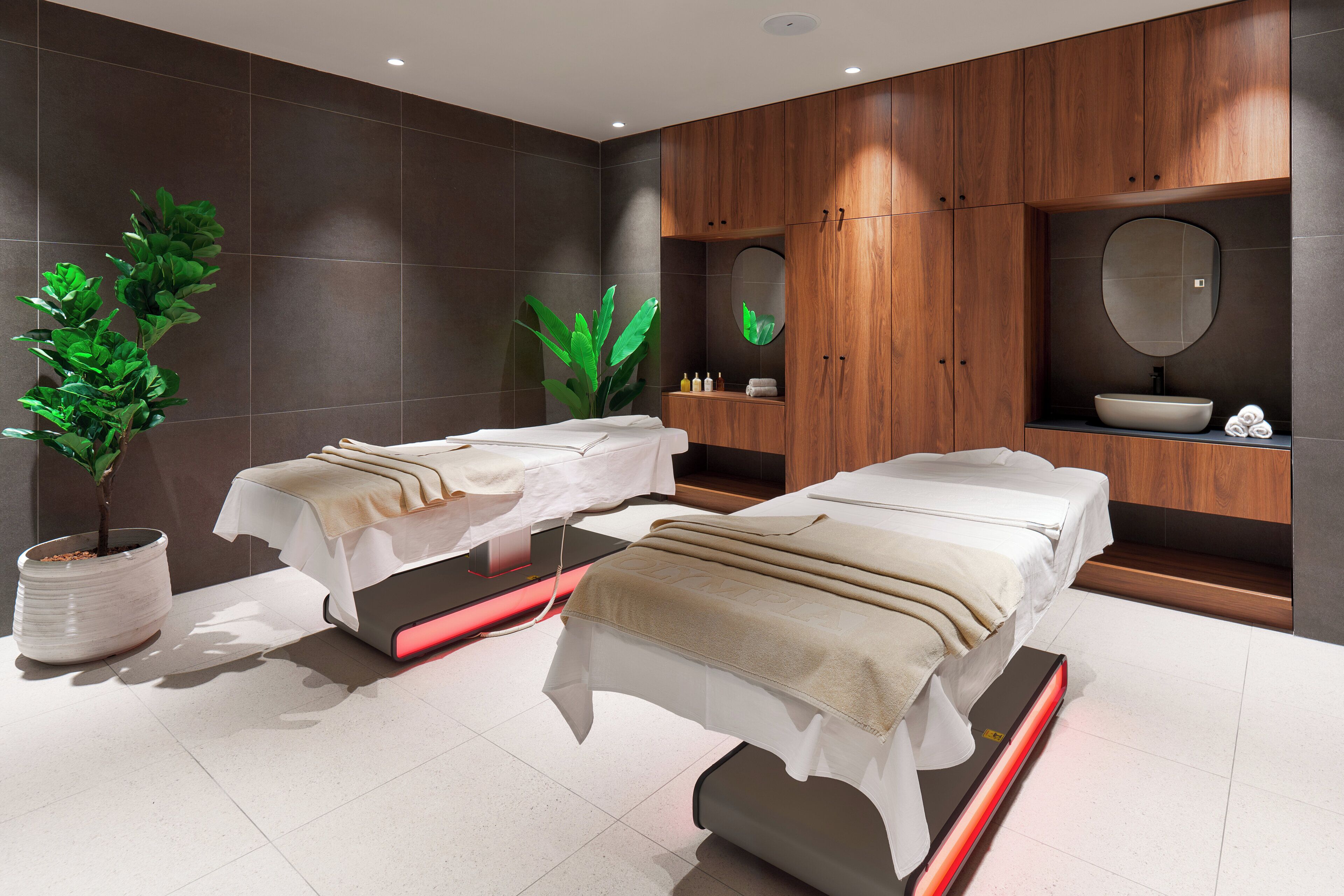 A modern spa room with two treatment beds, a large mirror, wooden cabinets, and potted plants.
