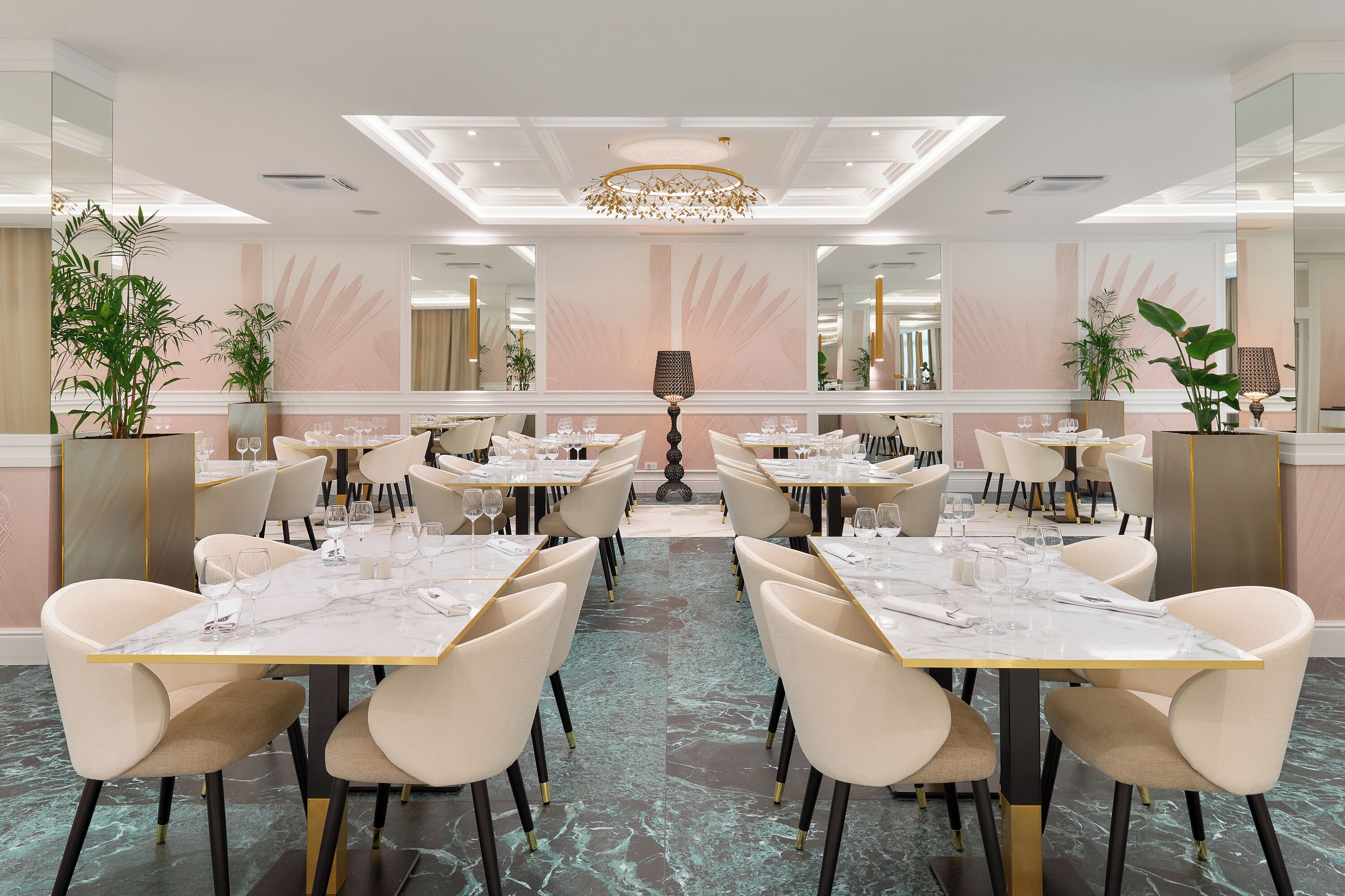 A spacious dining area with marble-topped tables and modern chairs, featuring a central chandelier and large mirrors on the walls.