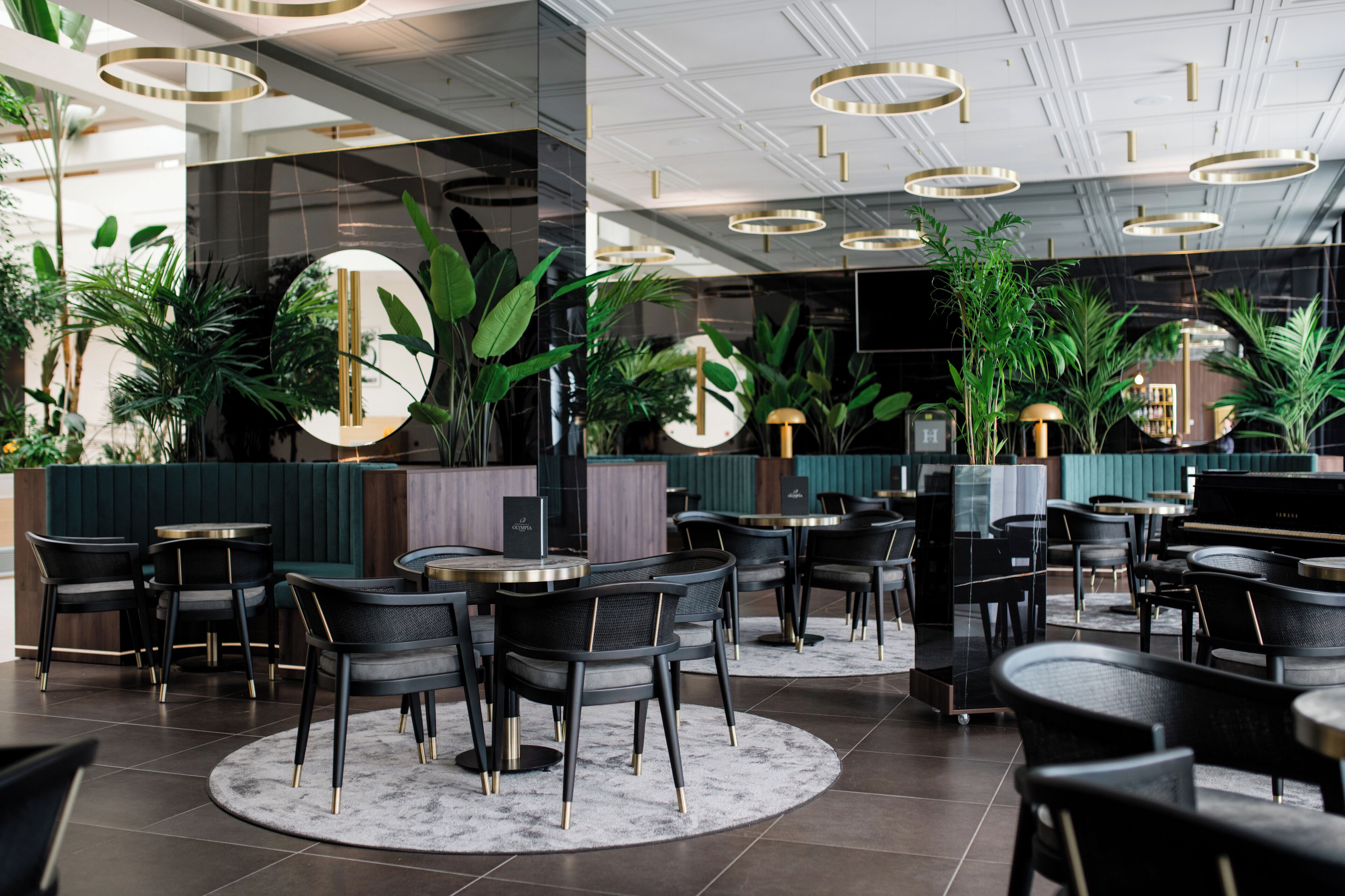 A modern indoor dining area with round tables and black chairs, surrounded by green plants and a piano.