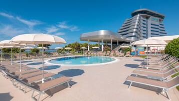 A poolside area with lounge chairs and umbrellas, a swimming pool, and a hotel building in the background.