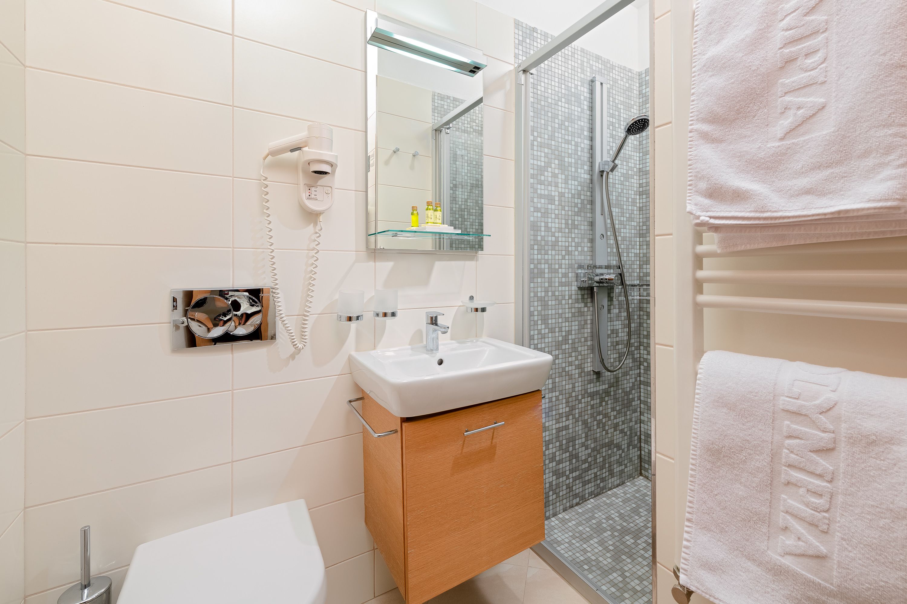 Un bagno moderno con un lavabo bianco, uno scaffale in vetro con articoli da toilette, un asciugacapelli e una zona doccia.