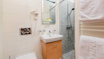 Un bagno moderno con un lavabo bianco, uno scaffale in vetro con articoli da toilette, un asciugacapelli e una zona doccia.