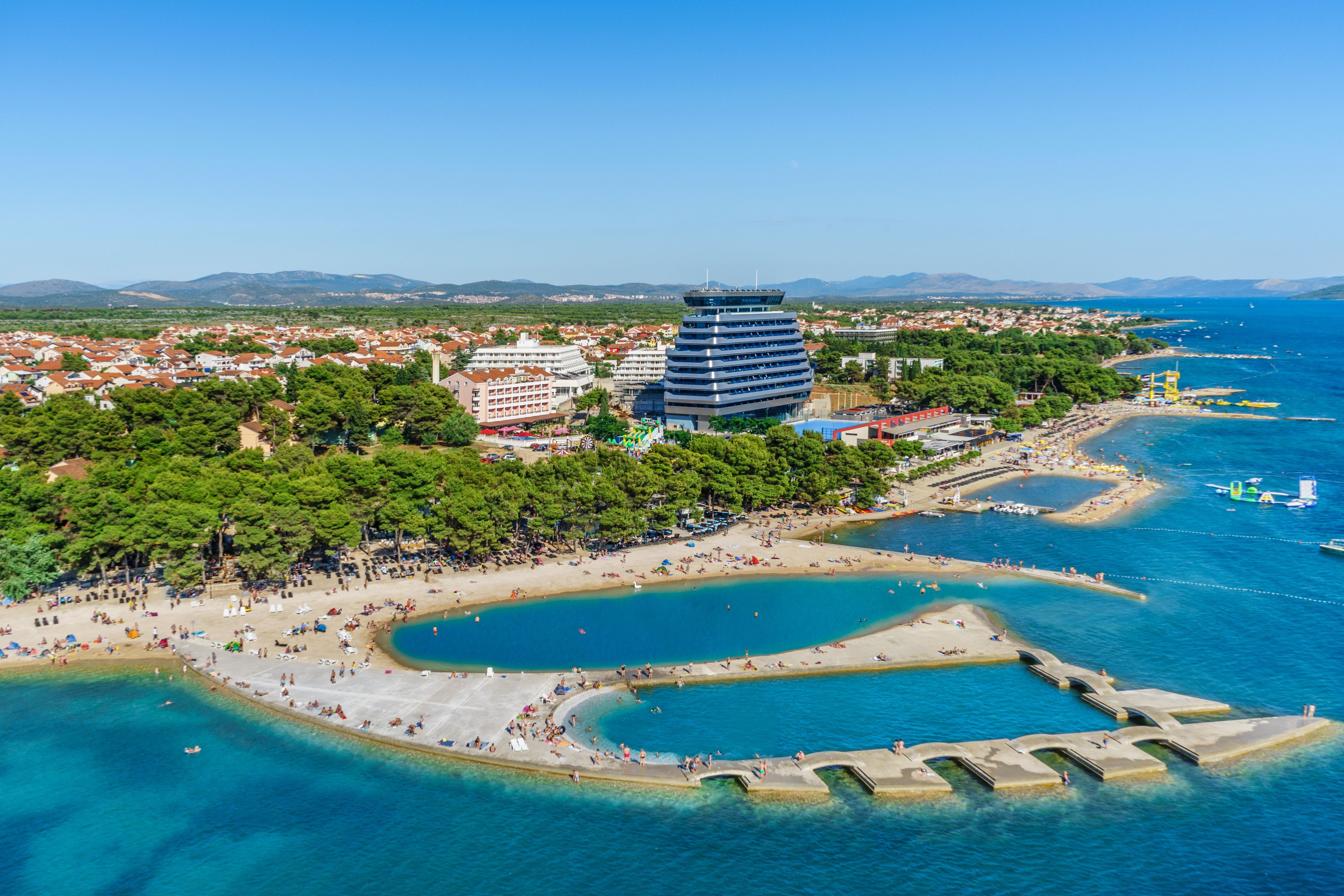 Vista aérea de una ciudad costera con un edificio moderno destacado, una playa y aguas azules cristalinas.