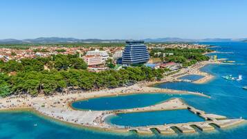 Vista aérea de una ciudad costera con un edificio moderno destacado, una playa y aguas azules cristalinas.