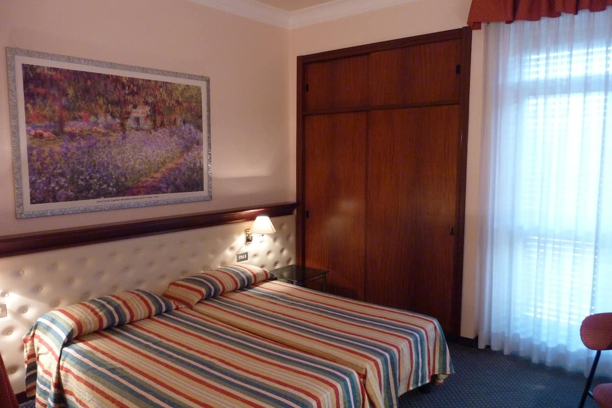 A hotel room with a large bed, a headboard with a light fixture, a wooden wardrobe, and a painting on the wall.