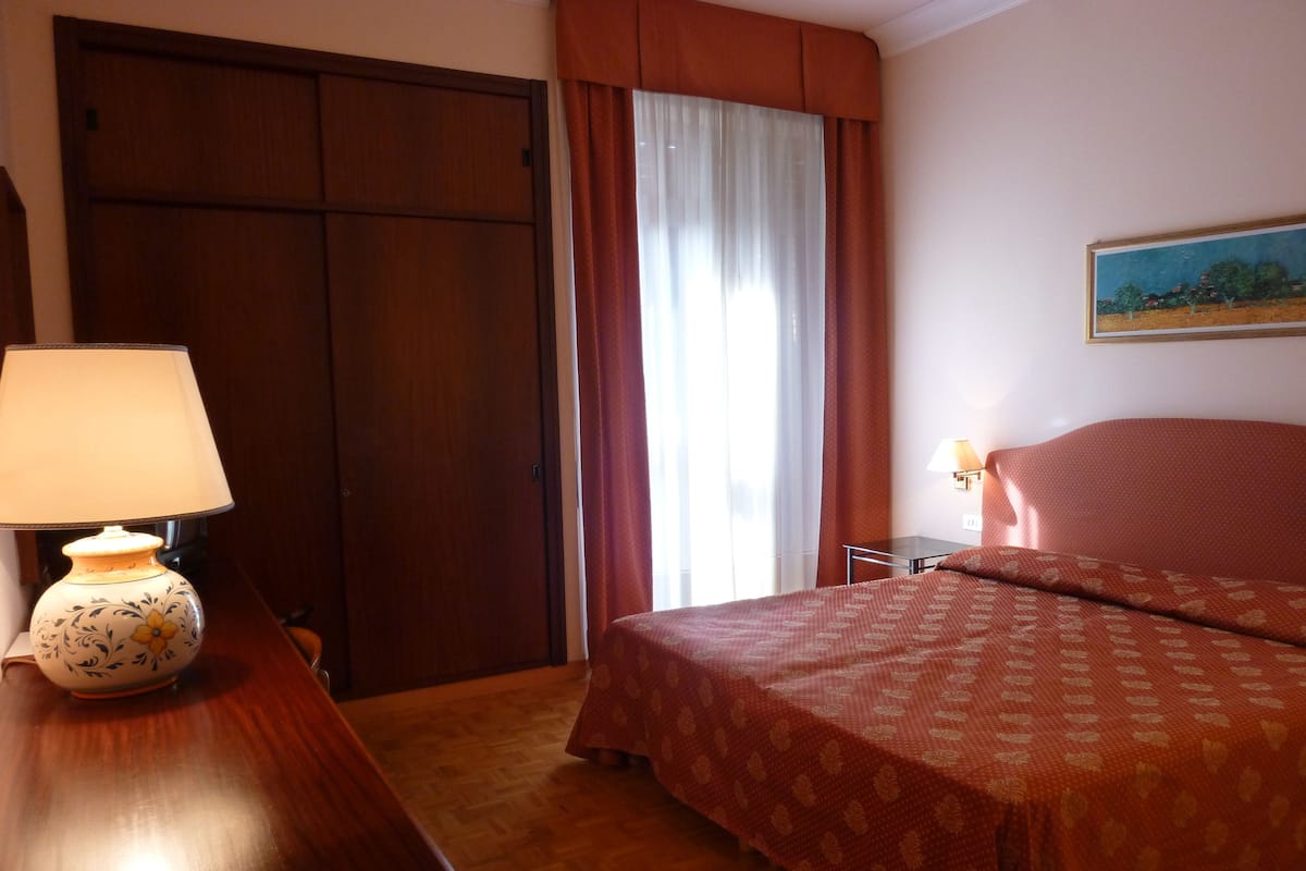 A hotel room with a wooden desk, a lamp, a bed with a patterned bedspread, a nightstand, and a painting on the wall.