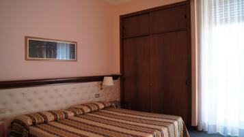 Habitación de hotel con una cama individual, un armario de madera, un cuadro en la pared y una ventana con cortinas de encaje.