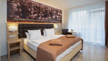 Habitación de hotel con una cama grande, cabecera, mesitas de noche, una pequeña mesa con lámpara y un ventanal con cortinas translúcidas.