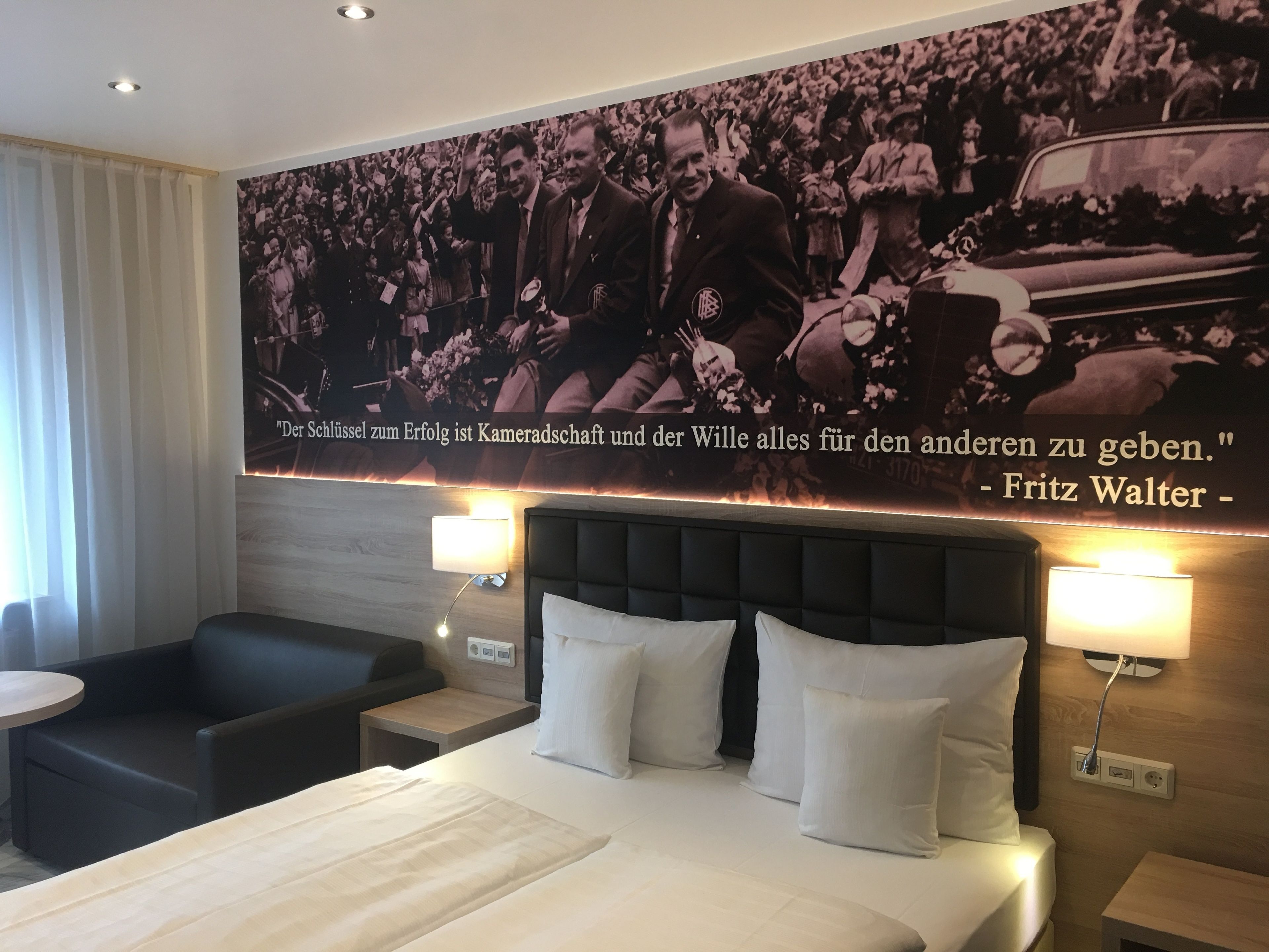 Een hotelkamer met een groot poster van een historisch gebeurtenis, een bed met wit beddengoed, twee nachtkastjes met lampen, en een donkergrijze bank.