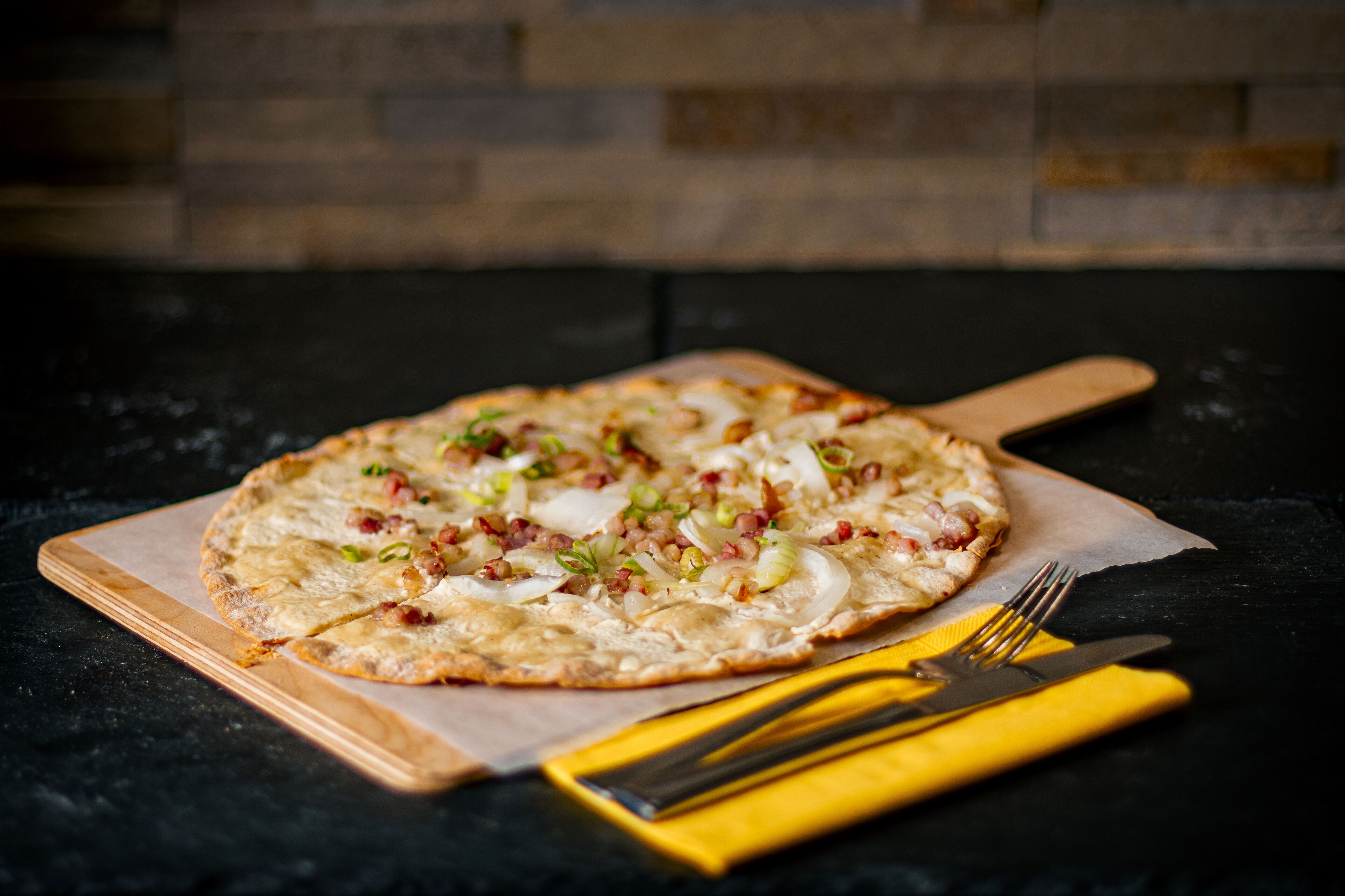 A pizza on a wooden board with a knife and fork on a yellow napkin.