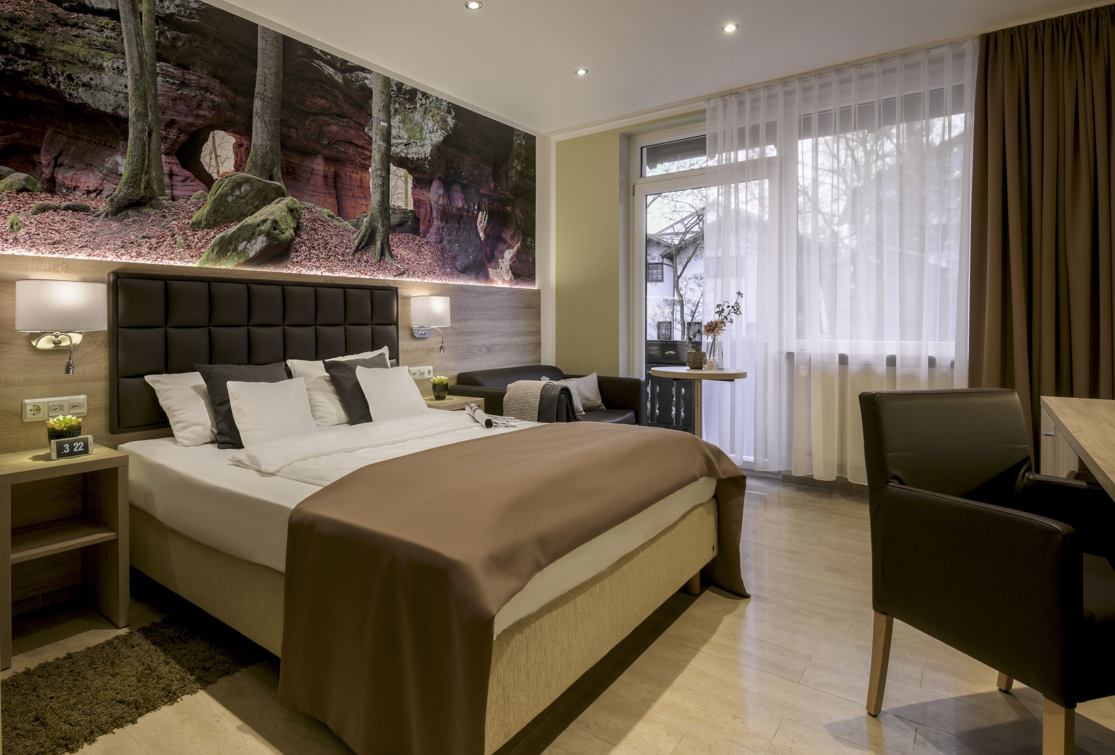 A modern hotel room with a large bed, a desk, and a view of trees through sheer curtains.