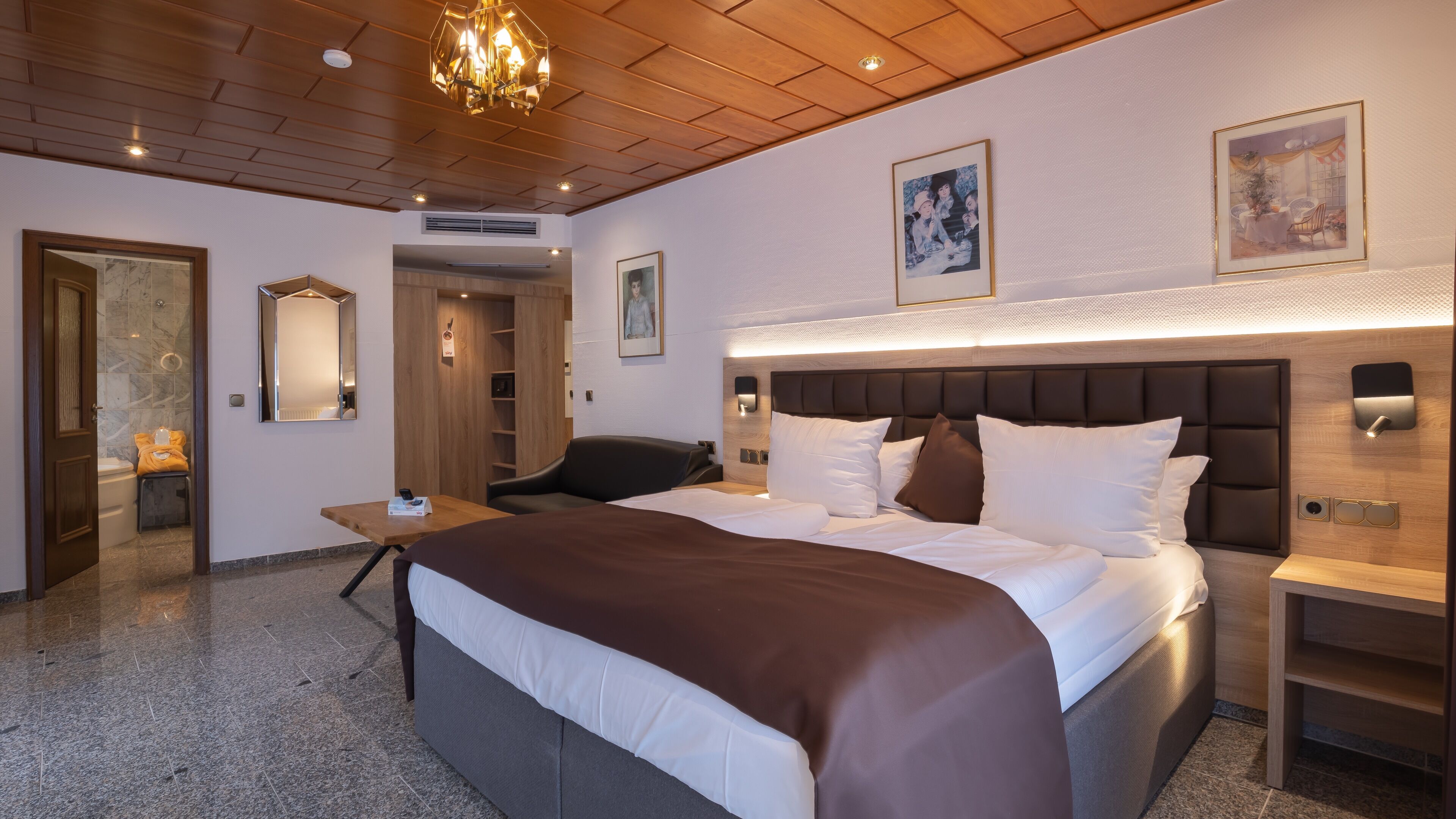 Une chambre d’hôtel avec un grand lit, une tête de lit en bois, une table de chevet, une chaise, un miroir et un lustre.