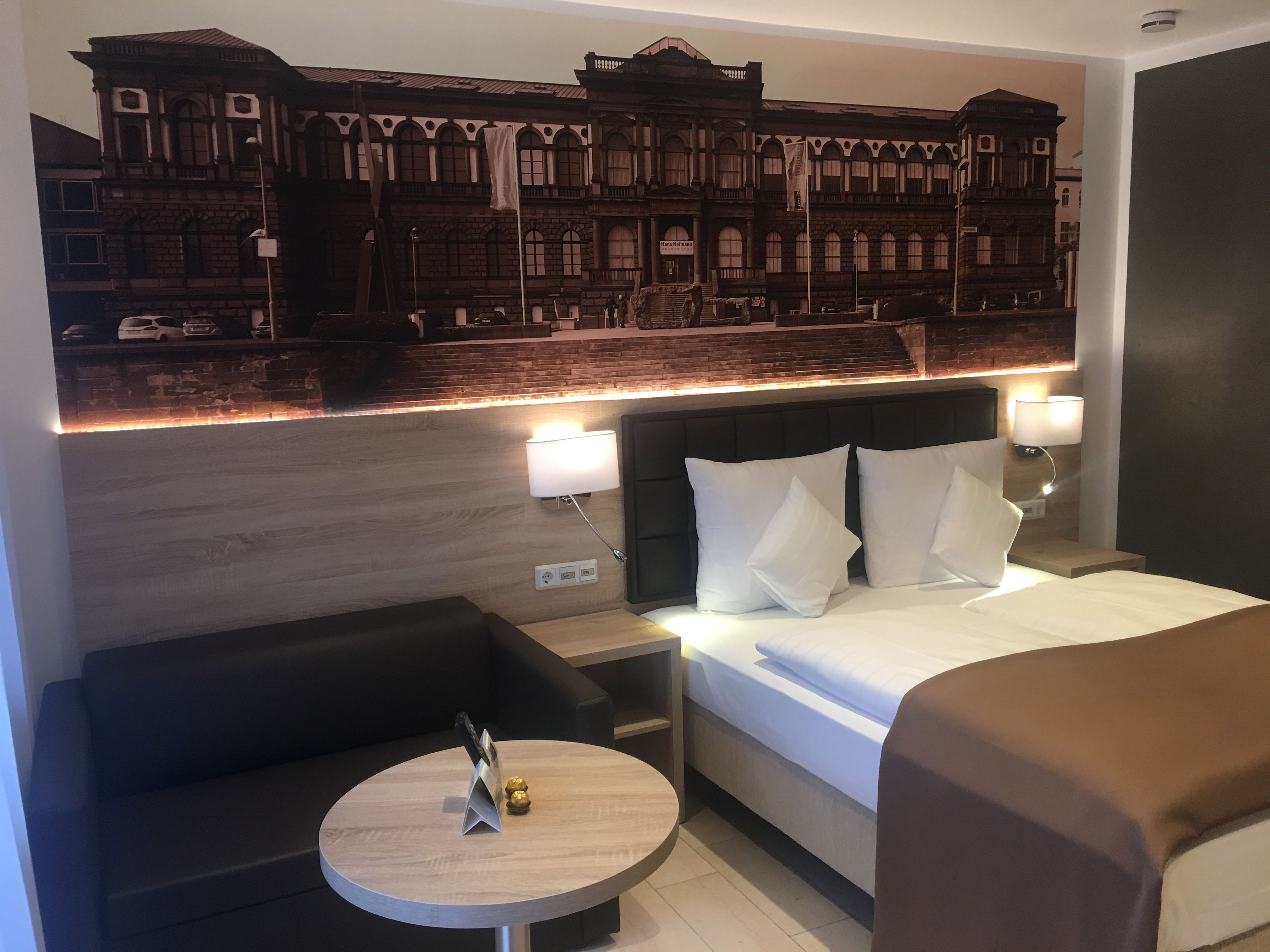 Habitación de hotel con una cama, dos mesitas de noche, una mesa redonda, un sofá y un mural grande en la pared que muestra un edificio.