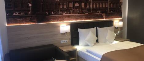 Habitación de hotel con una cama, dos mesitas de noche, una mesa redonda, un sofá y un mural grande en la pared que muestra un edificio.
