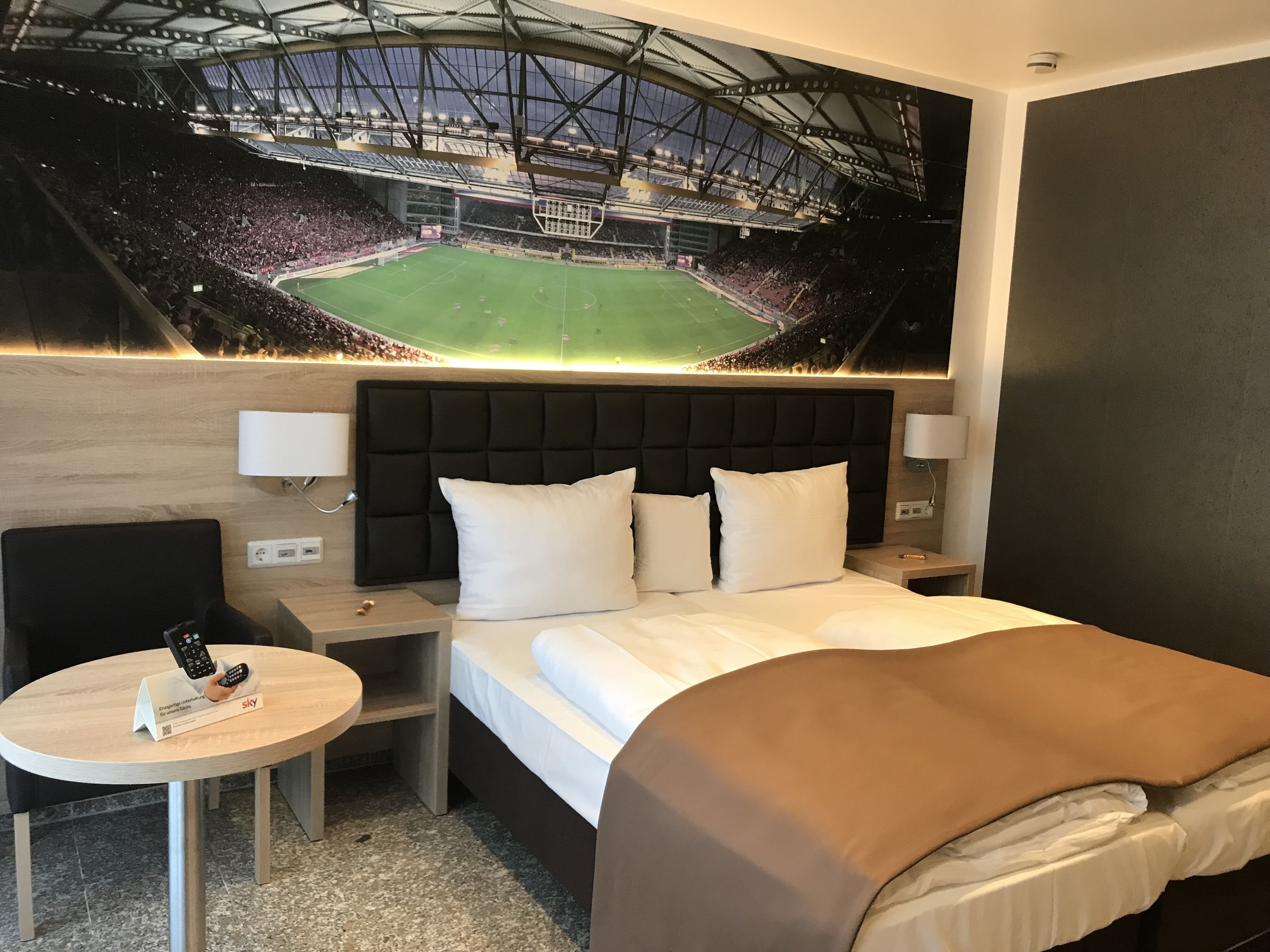 Una habitación de hotel con una cama, mesitas de noche, un escritorio y un mural grande de un estadio.