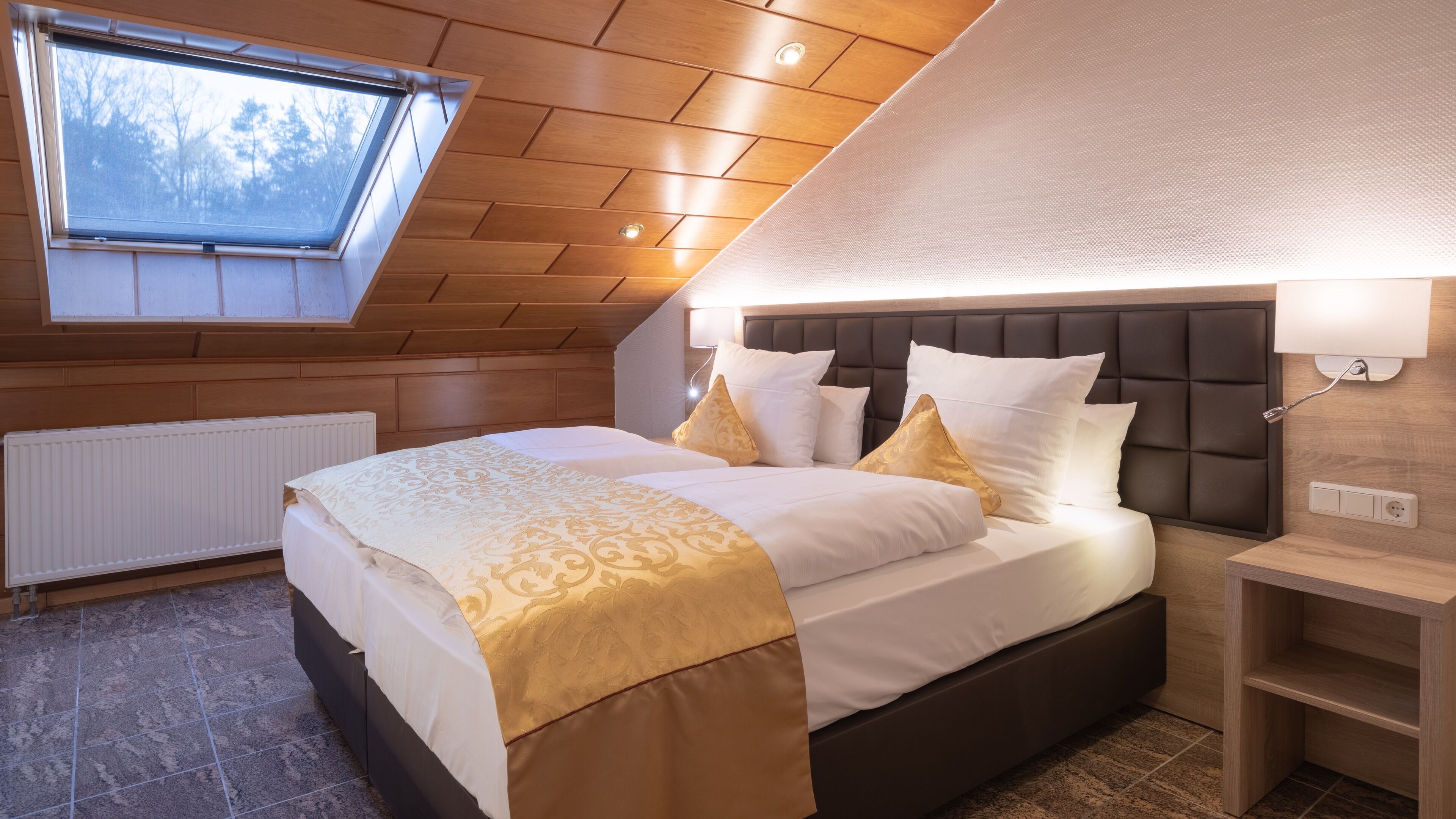 Ein Schlafzimmer mit schräg angeschnittener Decke, einem großen Bett mit weißer und goldener Bettwäsche, einem Kopfteil und einem Fenster.