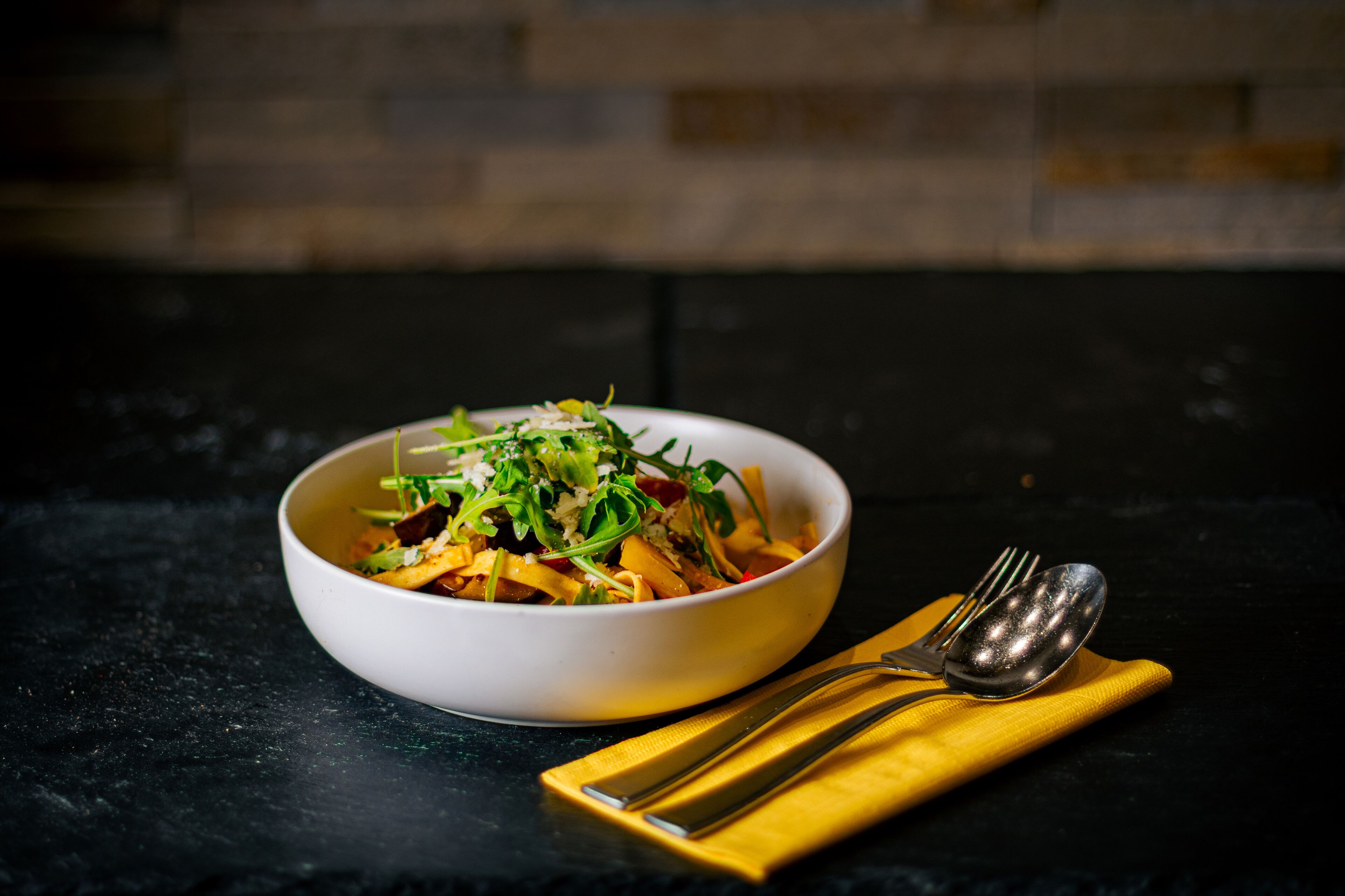 Un plato de pasta con verduras y queso sobre una superficie oscura, con cuchara y tenedor sobre una servilleta amarilla.