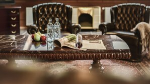 Une table en marbre, avec un journal, deux bouteilles et des pommes. Deux fauteuils en cuir sont placés derrière la table.