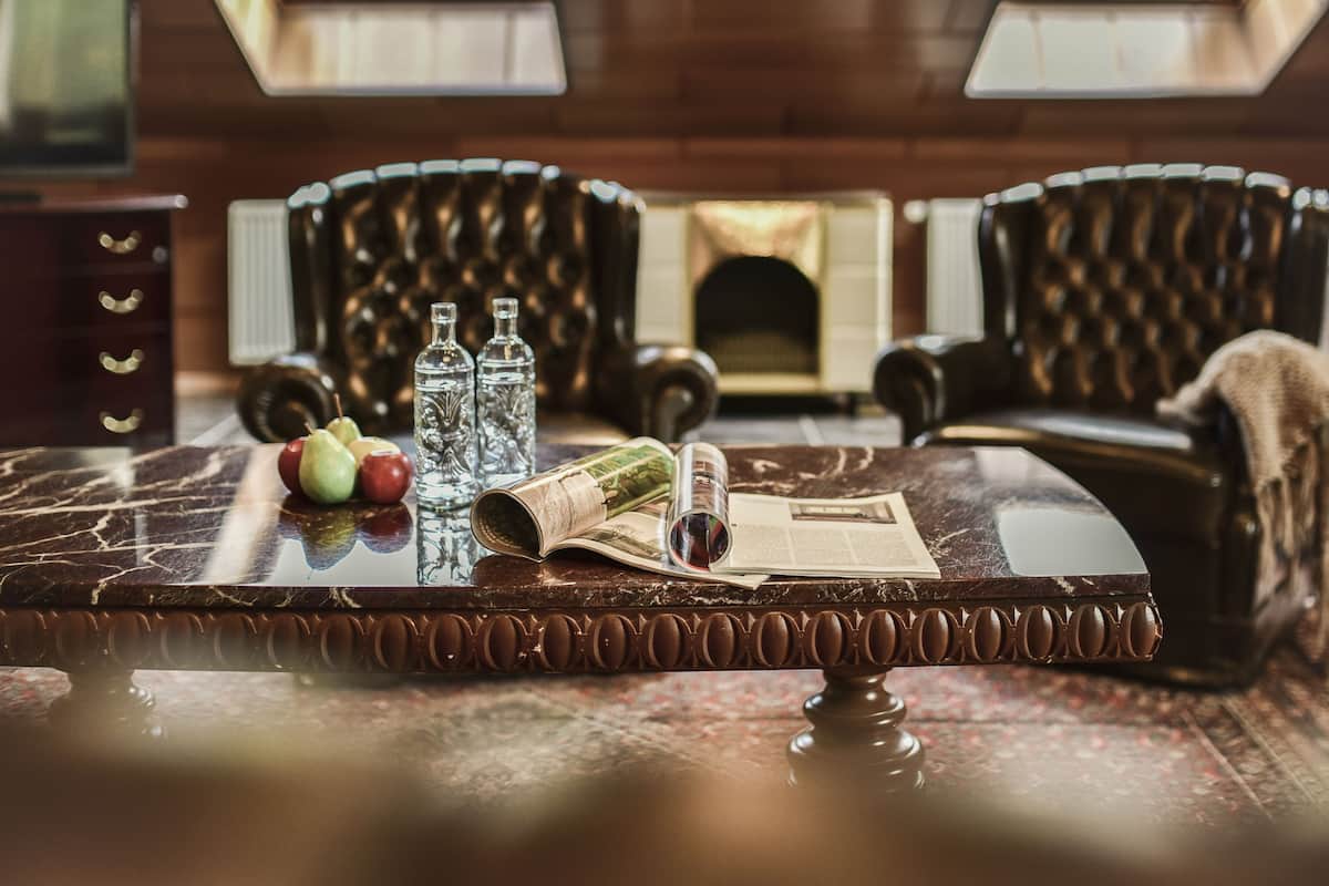 Una mesa de mármol con un periódico, dos botellas y manzanas. Dos sillones de cuero están situados detrás de la mesa.