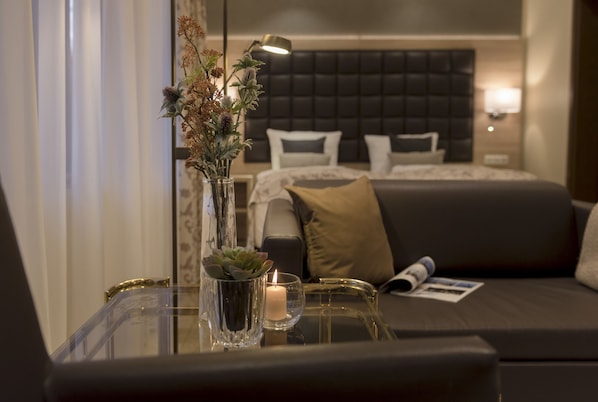 Una habitación de hotel moderna con una mesa de vidrio, un florero con flores secas, una vela encendida y una cama con cabecera oscura y muchos almohadones.