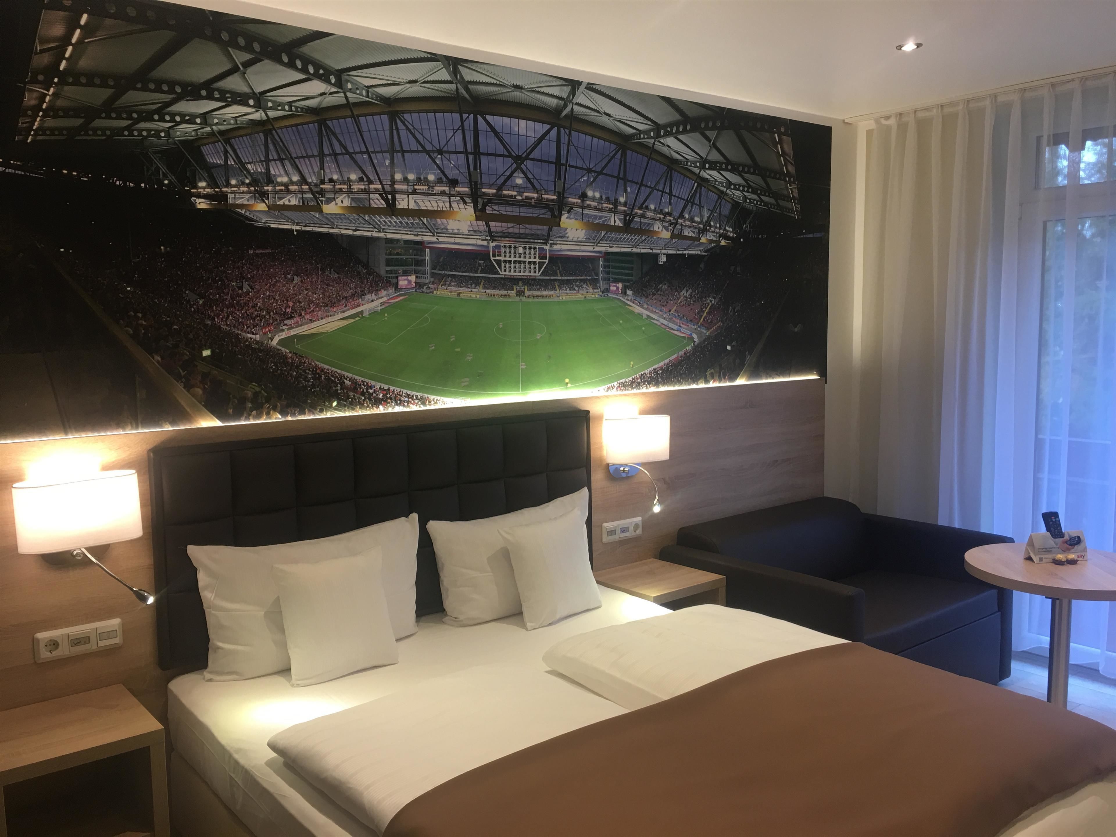 Habitación de hotel con cama, mesita de noche, lámpara, una pequeña mesa redonda y un cuadro de un estadio.