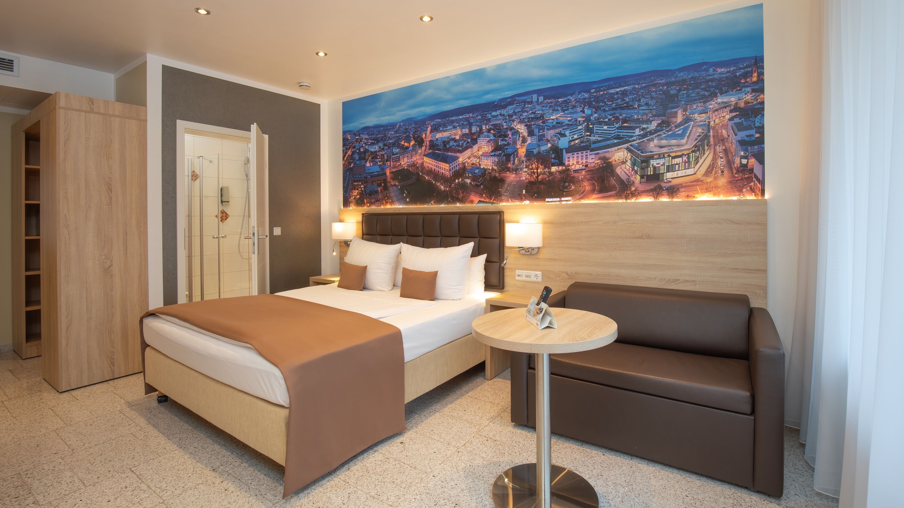 Une chambre d’hôtel avec un lit, un canapé, une petite table, une douche et une fresque murale représentant un paysage urbain.