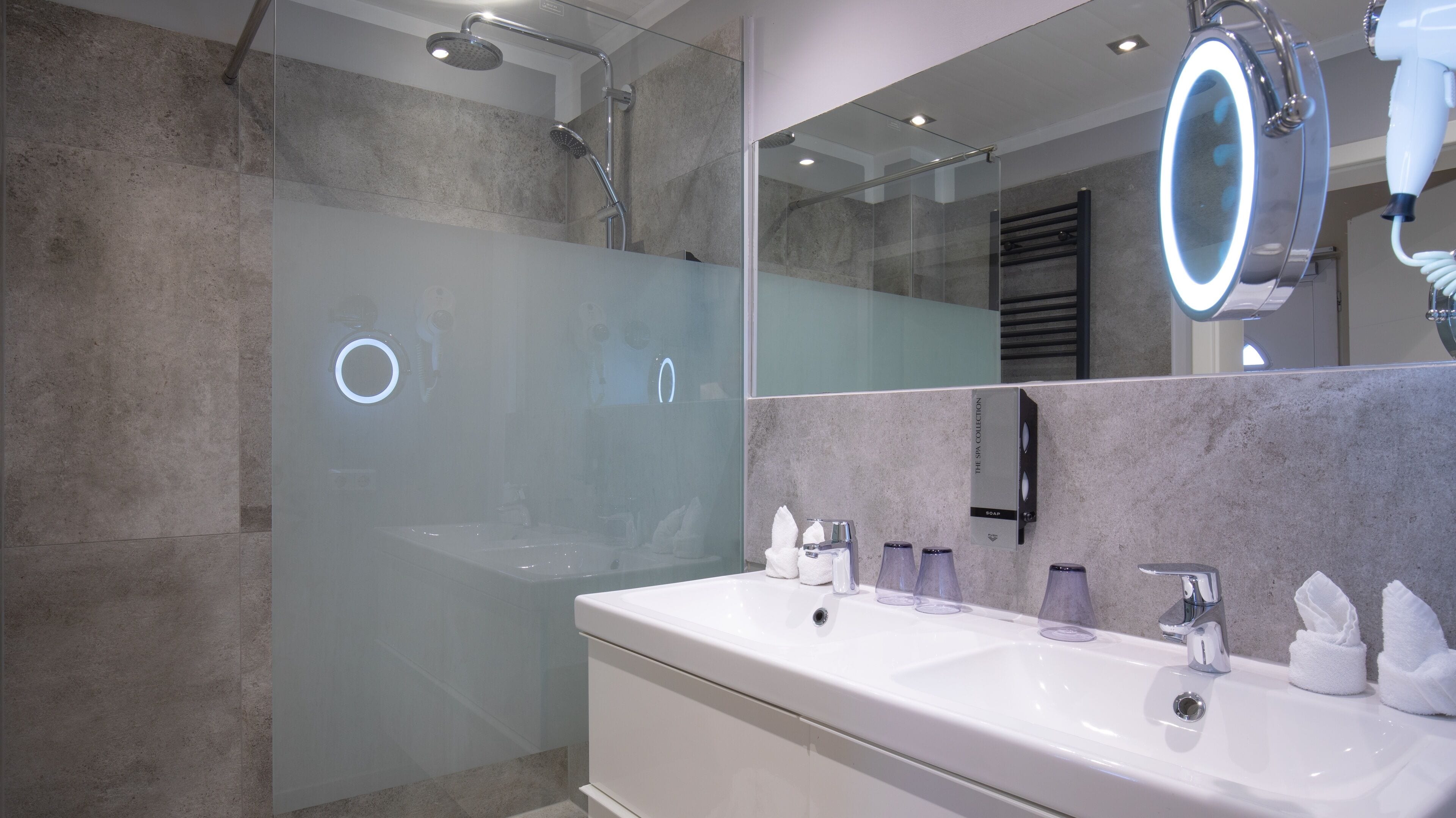A modern bathroom with a glass-enclosed shower, a large mirror with a built-in light, and a white vanity with multiple sinks and towel dispensers.