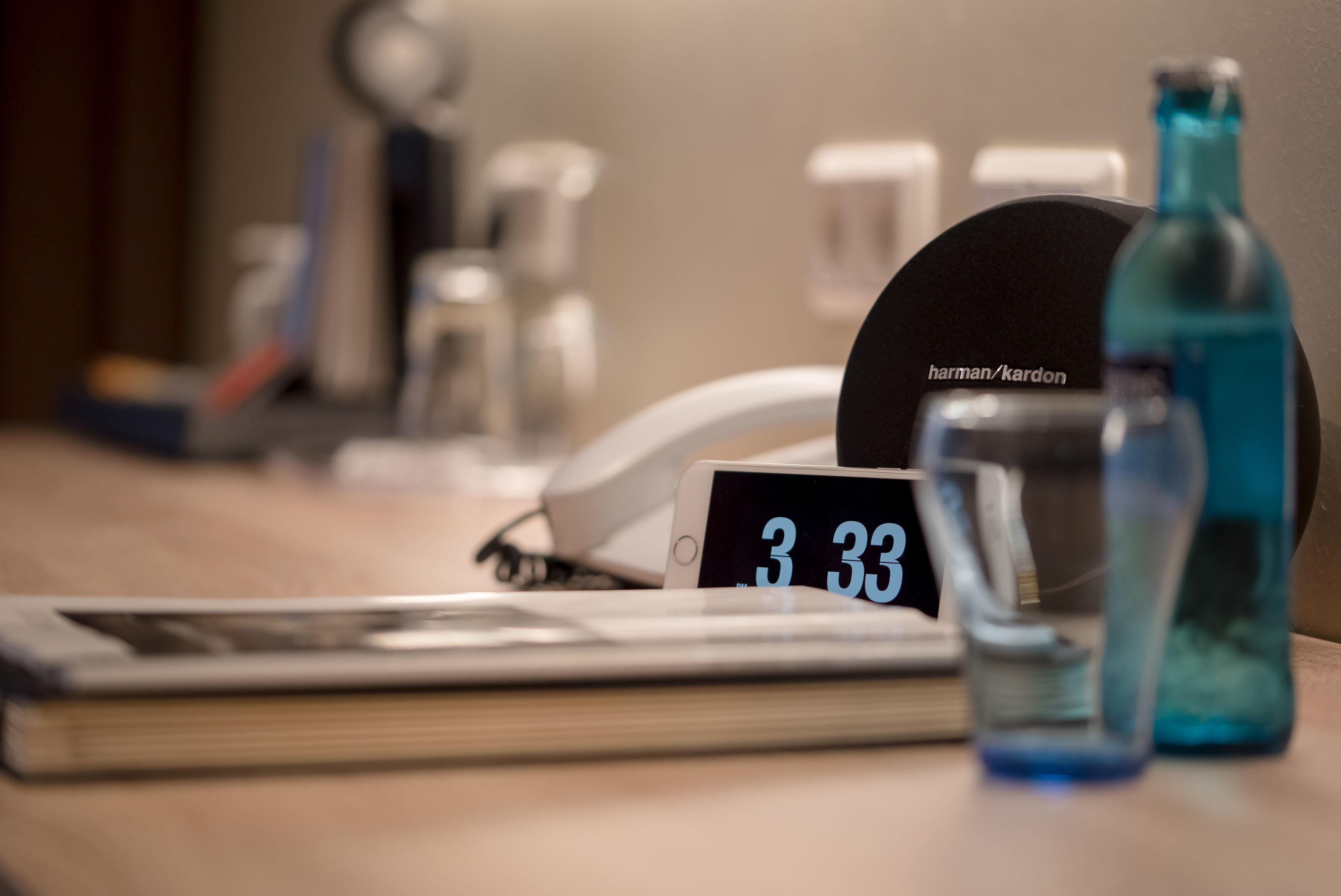 A desk with a digital clock displaying 3:33, a glass of water, a bottle, and a phone.