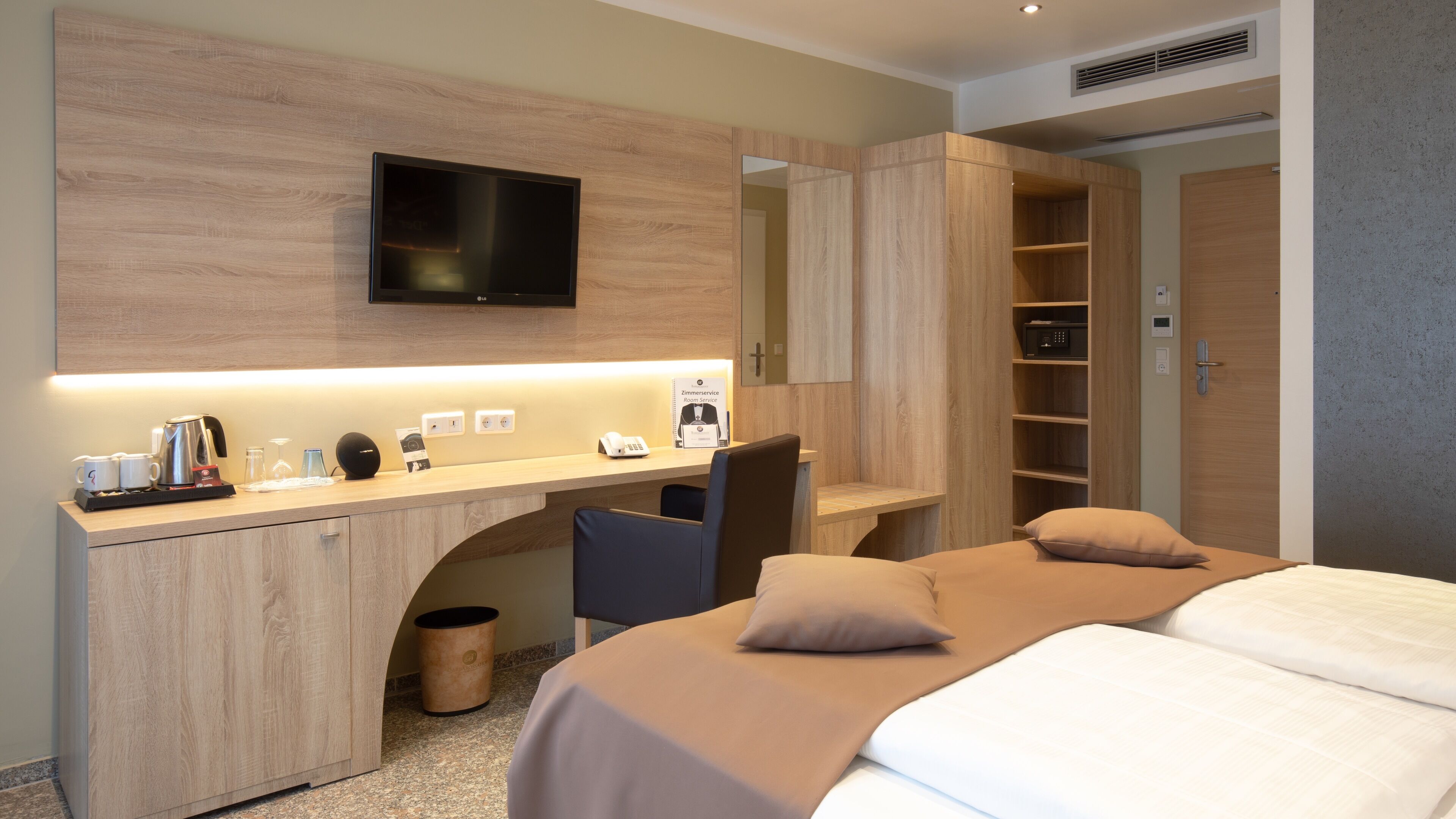 A modern hotel room with a wooden desk, a flat-screen TV, a coffee maker, and a bed with beige bedding.