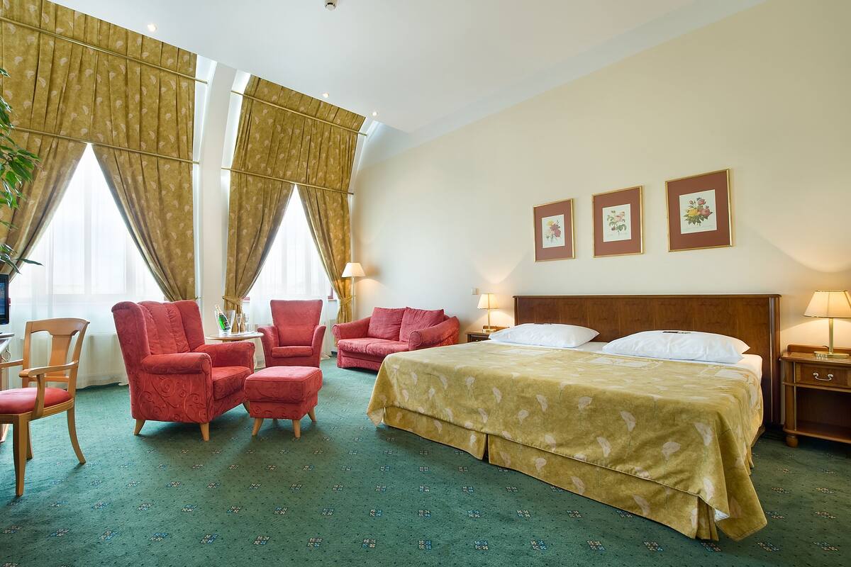 Habitación de hotel con una cama grande, dos sillones rojos, una silla de madera, una mesita con una botella de champán y una lámpara.