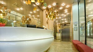 A reception area with a large white reception desk, a red bench, and an elevator. - EA Hotel Rokoko (Prague)