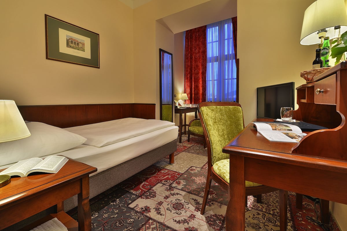 Une chambre d’hôtel avec un lit, un bureau, une chaise, une lampe, un cadre photo et une fenêtre avec des rideaux.