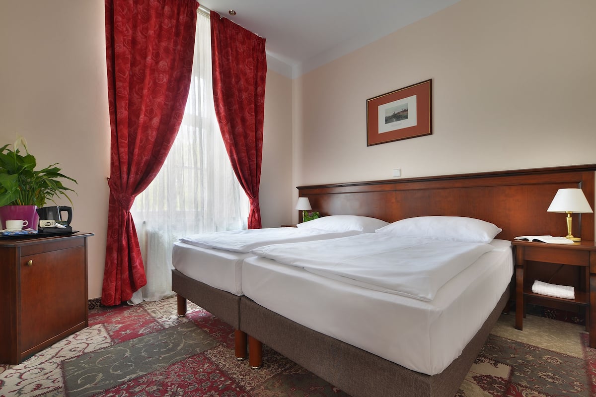 Une chambre d’hôtel avec deux lits, une tête de lit en bois, une table de chevet avec une lampe, un bureau avec une plante et un tableau encadré au mur.