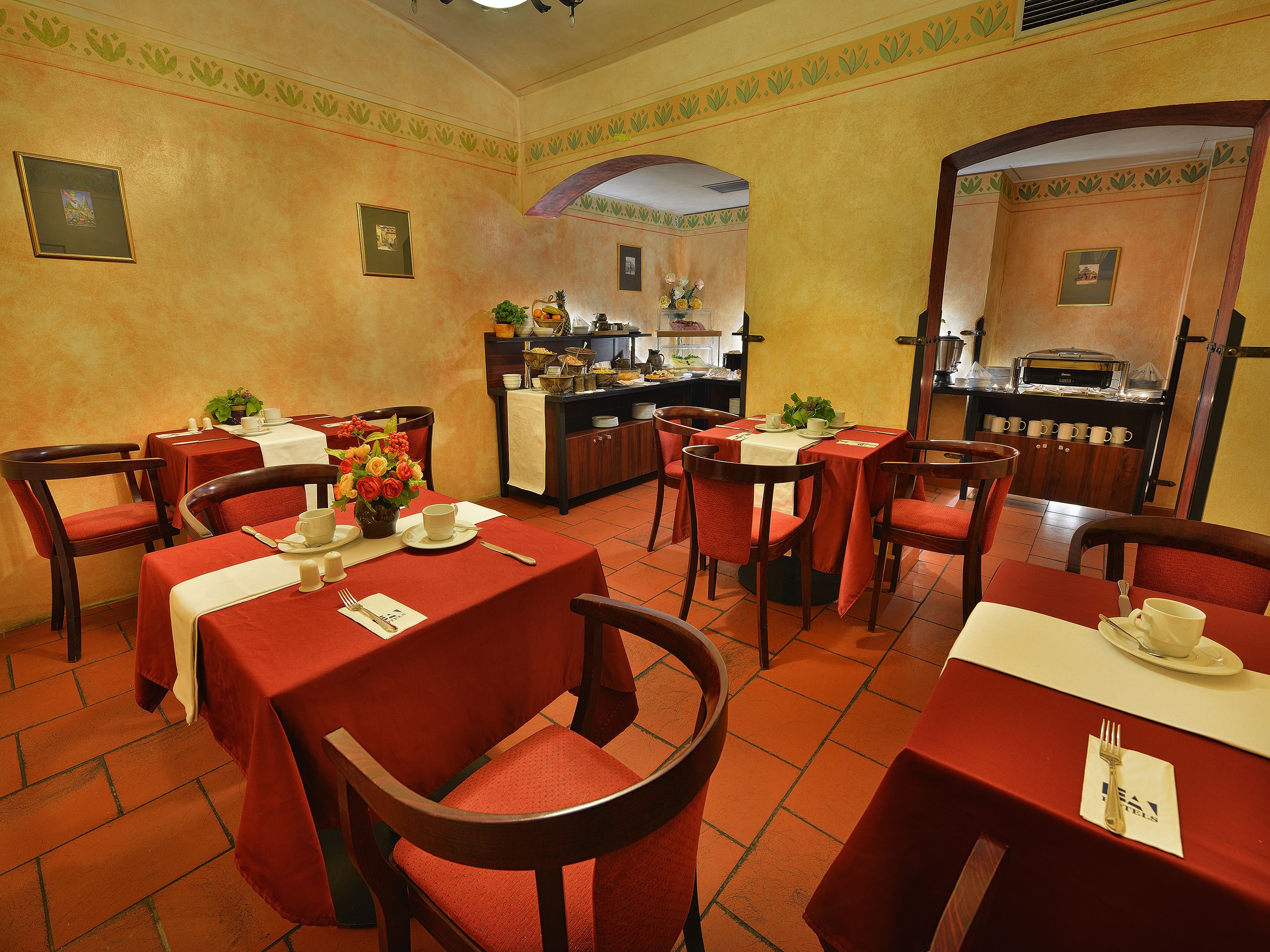 A restaurant with red tablecloths, wooden chairs, and a kitchen area in the background.