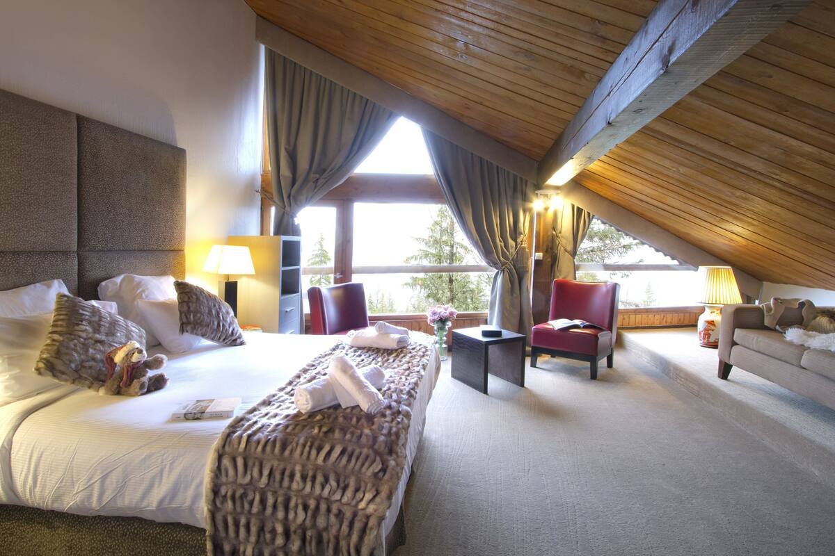A spacious bedroom with a large bed, a red armchair, a sofa, and a wooden ceiling.