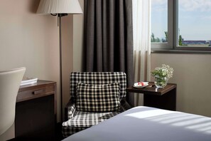 A hotel room with a bed, a chair, a nightstand, a lamp, a vase with flowers, and a window with a view.