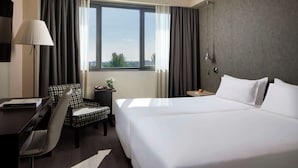 A hotel room with a large bed, a desk with a chair, a lamp, and a view of the outdoors through a window. - NH Alcorcón (Alcorcon)