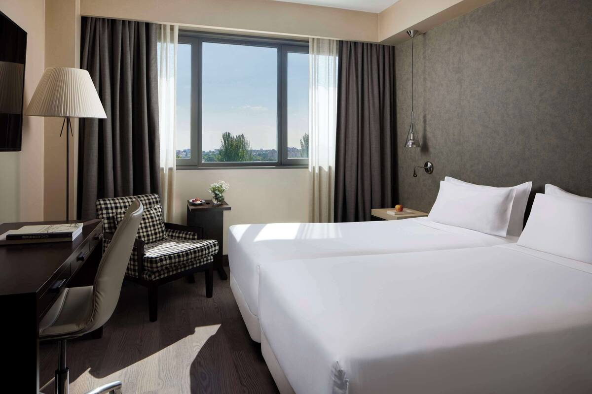 A hotel room with a large bed, a desk with a chair, a lamp, and a view of the outdoors through a window.